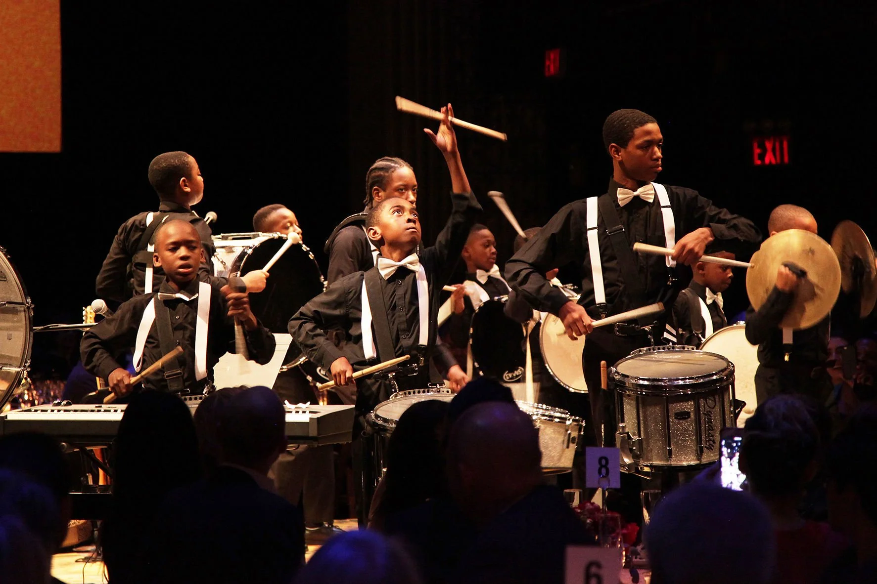 A group of young boys dressed in black shirts and bow ties performing on stage with drums and cymbals, with some playing drums and others with percussion instruments, under stage lights in front of a dark background.