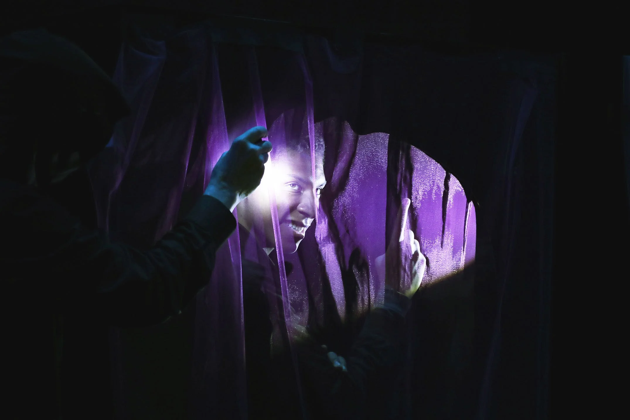 A man is pulling back a purple curtain to reveal a bright digital display of a smiling woman's face.