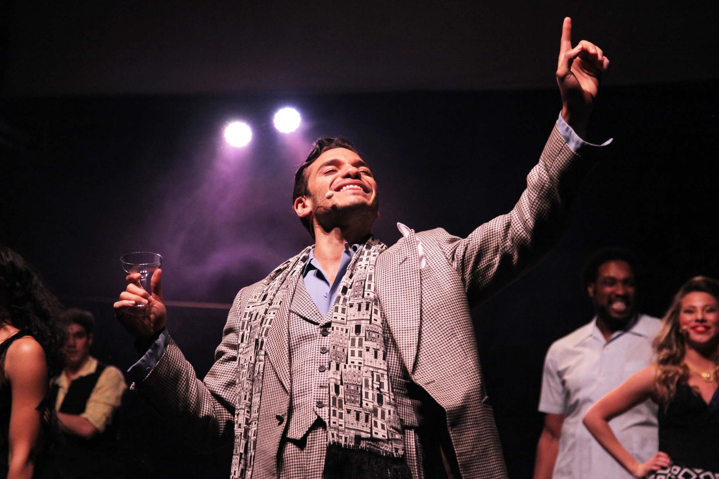 A man in a patterned suit is smiling with his eyes closed, raising one hand with his index finger points up, at a social event or celebration. Several people are around him, some smiling, in a dark venue with stage lights.