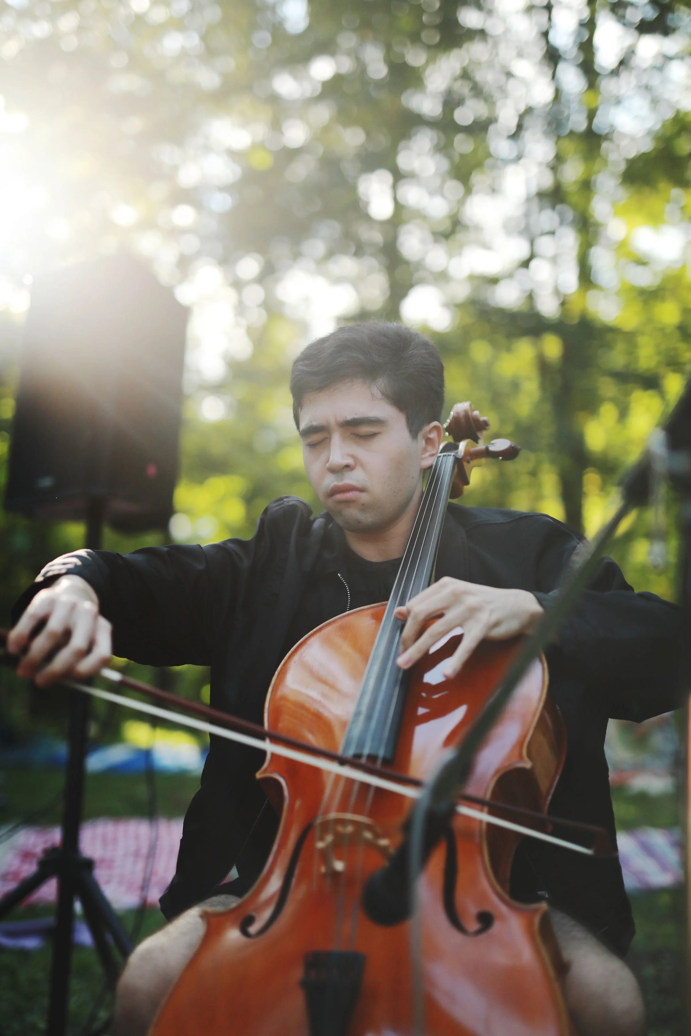 Young man with dark hair playing a cello outdoors during daylight, surrounded by trees with sunlight filtering through the leaves.