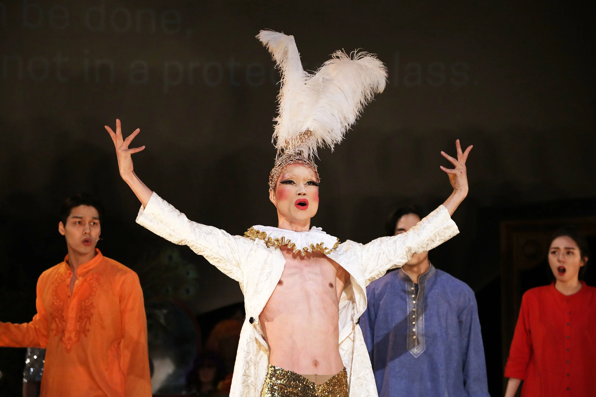 A performer in elaborate costume with a large feathered headpiece and gold sequined pants, singing or speaking with arms raised, flanked by supporting performers in colorful traditional costumes.