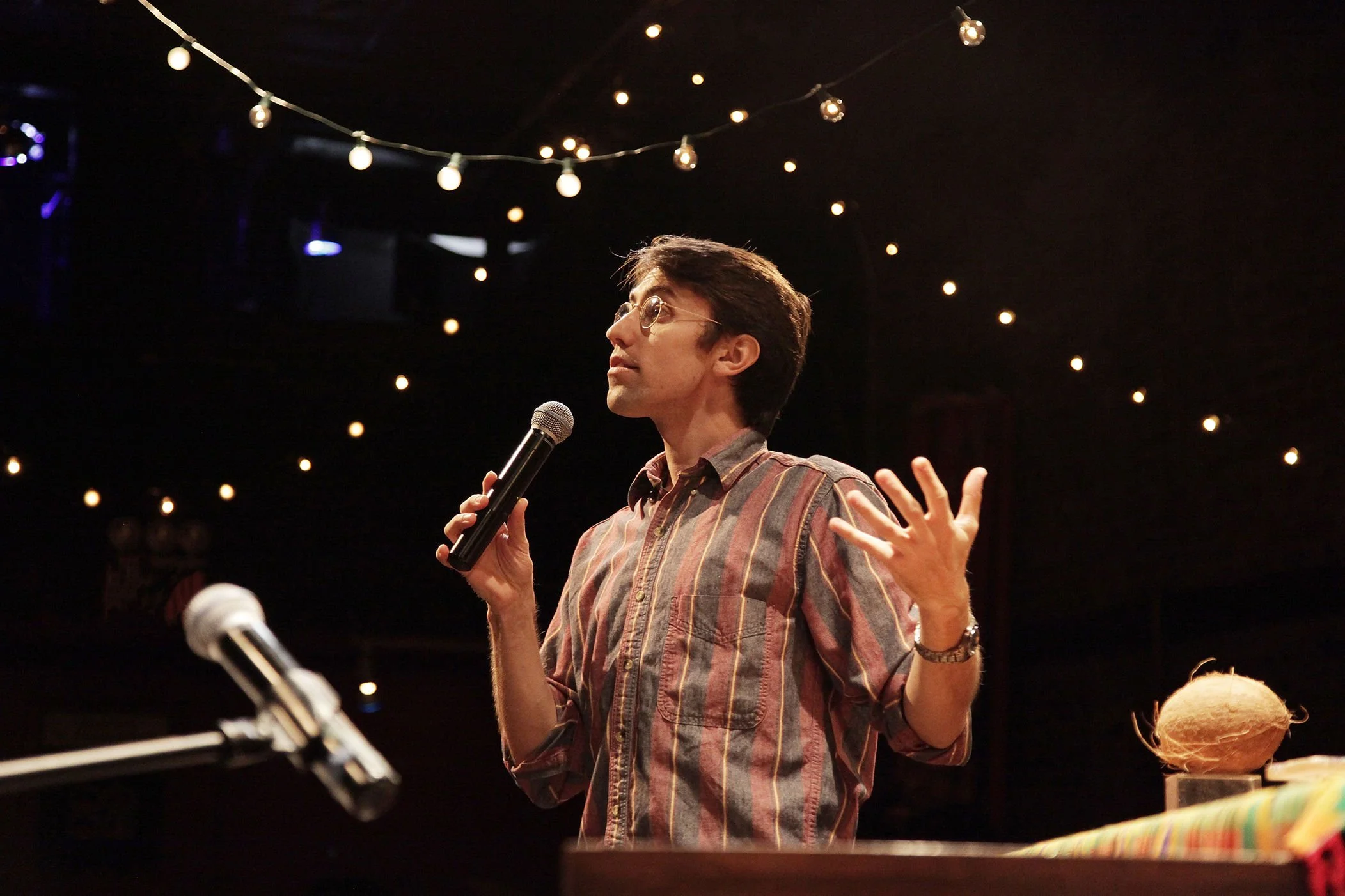 A man with glasses, wearing a plaid shirt, holding a microphone, speaking at an indoor event with string lights overhead.
