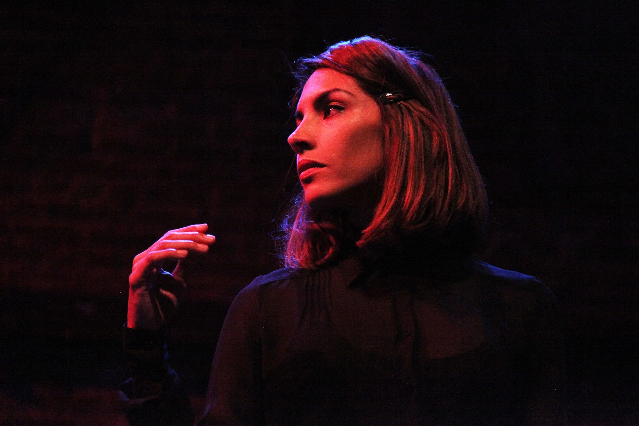 A woman with long, reddish hair and a black top, holding her hand near her face, is illuminated by red and blue stage lighting against a dark background.