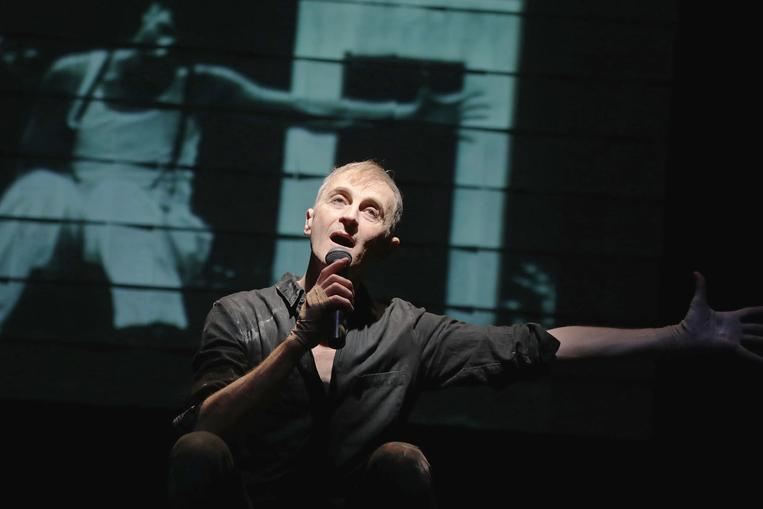 Man speaking and gesturing with one hand while holding a microphone with the other, in a dimly lit setting with a building's reflection in the window behind him.