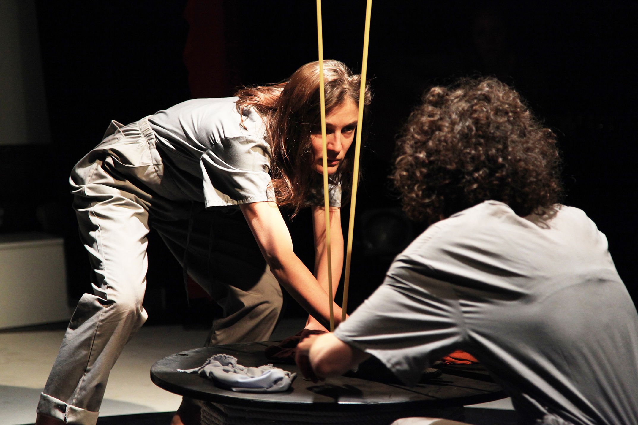 Two women in gray scrubs participating in a theatrical performance, with one woman bending over a black table and the other woman crouching on the floor, set against a dark background.