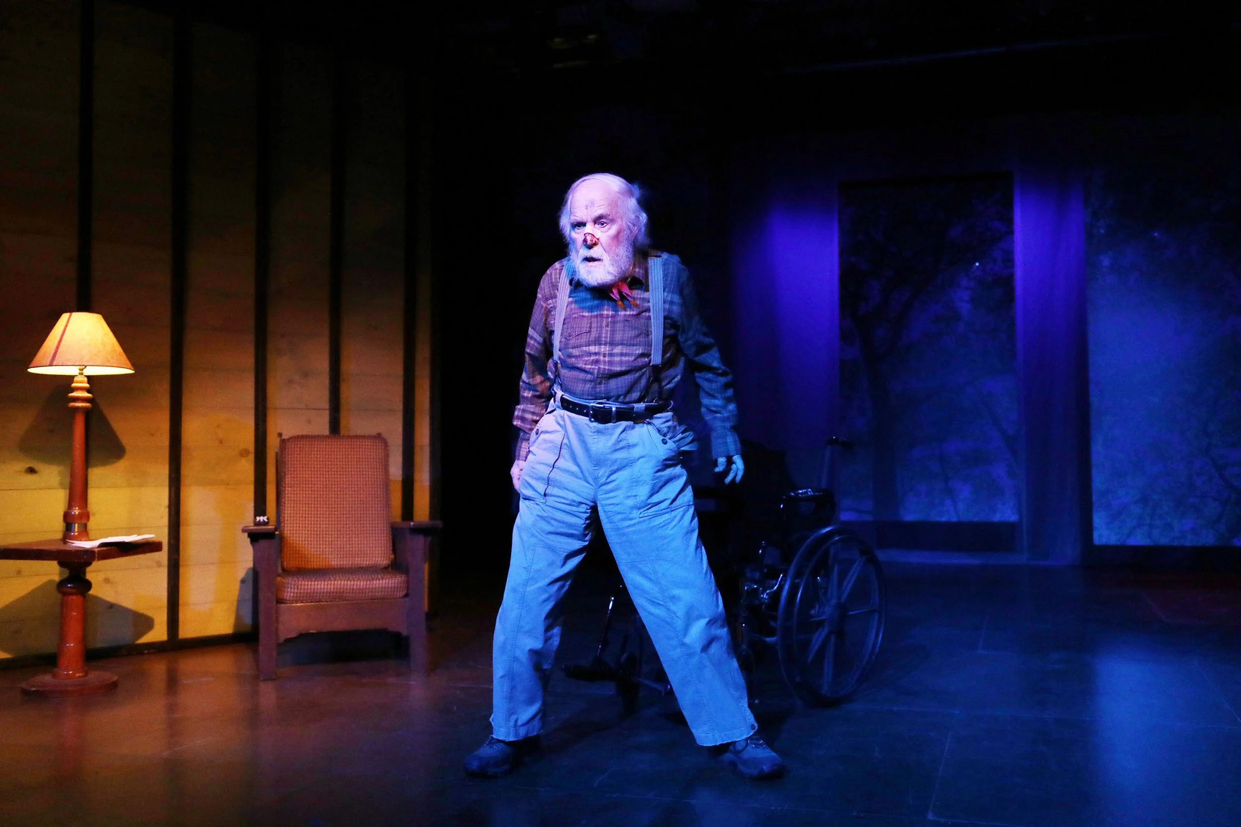 A stage scene with an elderly man in a plaid shirt, suspenders, and light-colored pants, with a bandaged nose, standing and looking surprised or intense. The stage has a wooden wall, a lamp, an armchair, and a wheelchair, with blue and warm lighting.