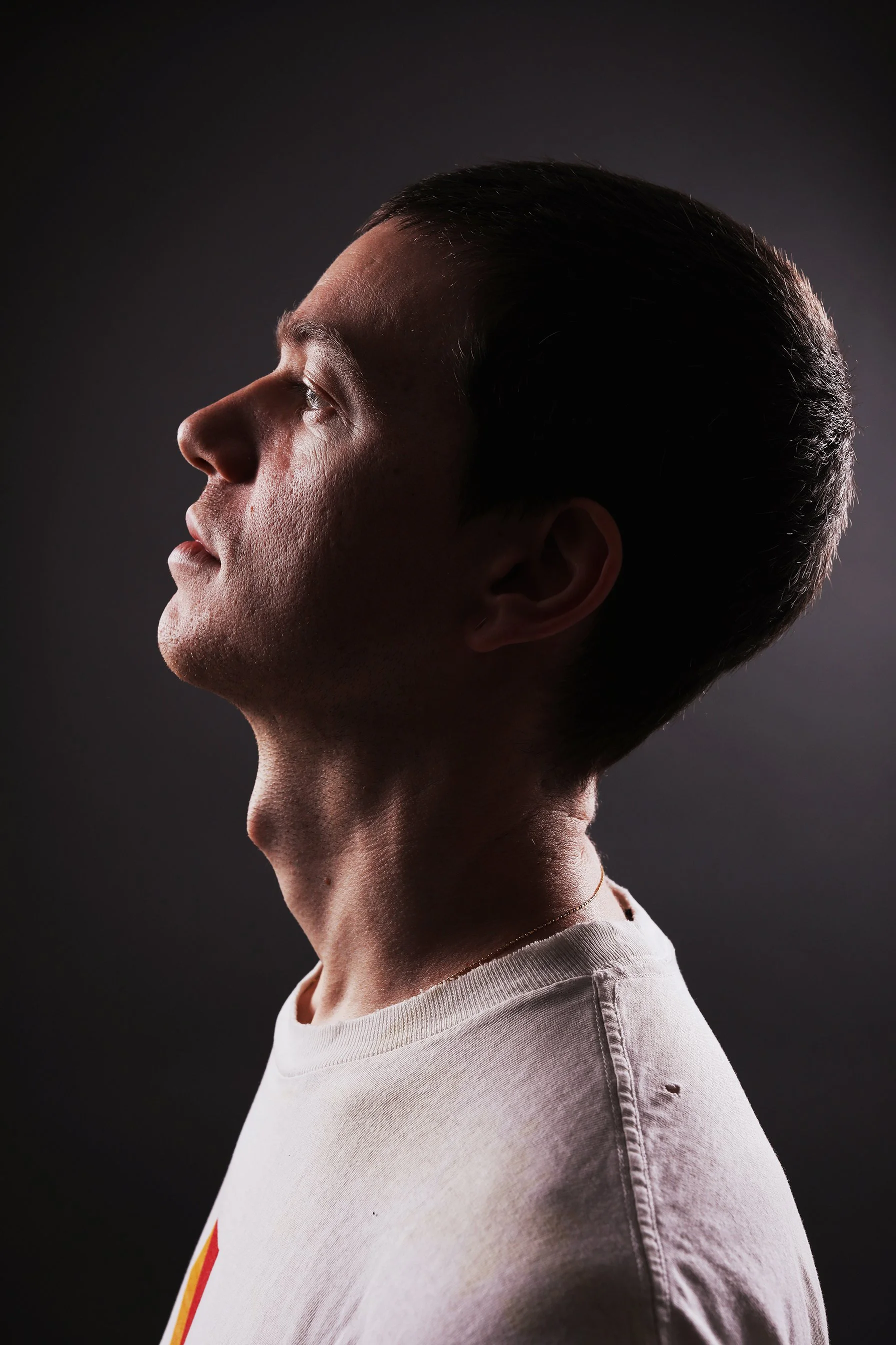 A side profile of a young man with dark hair, wearing a white T-shirt with a colorful stripe on the side, against a dark background.