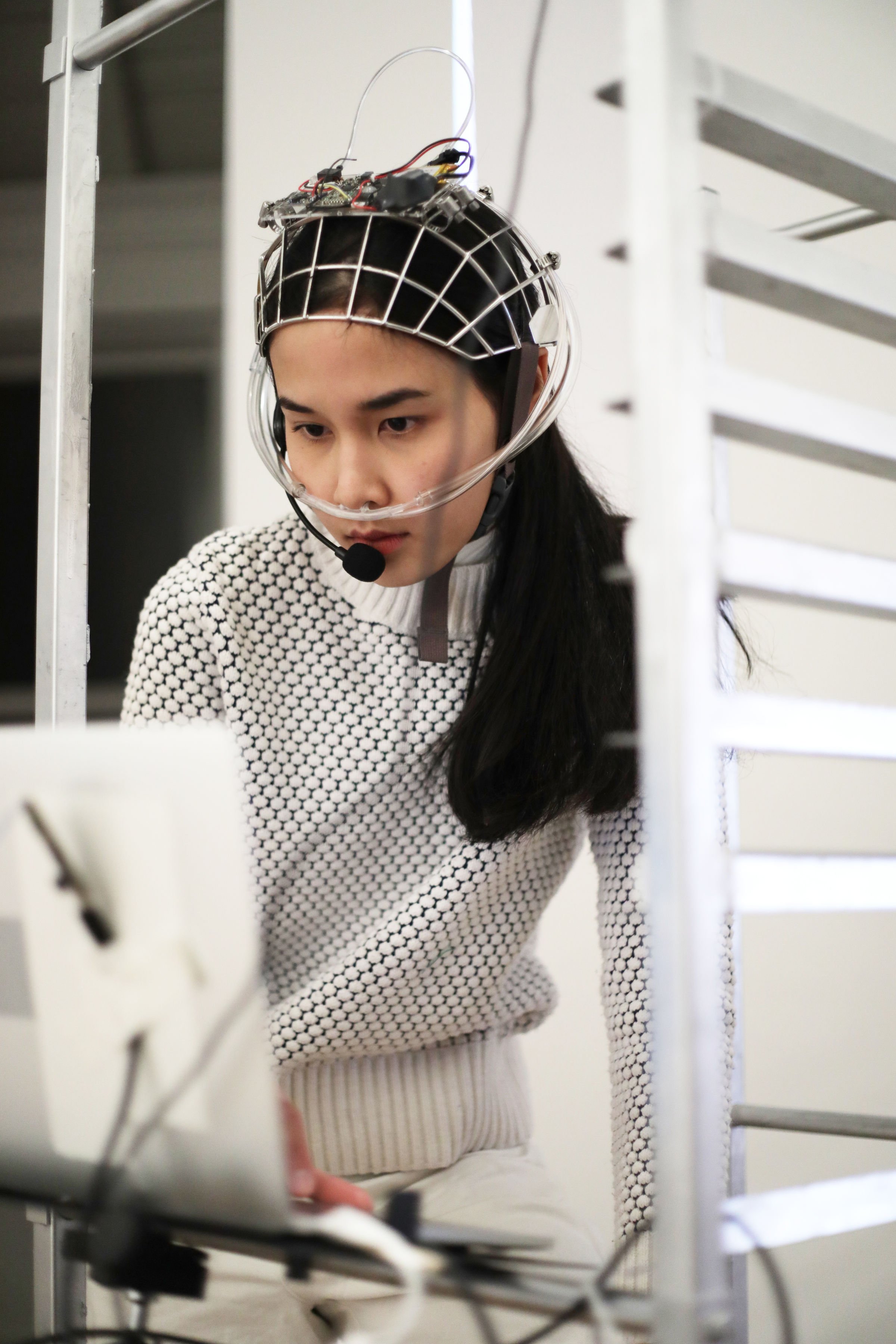 A woman wearing a transparent helmet with wires and electronic components, possibly a virtual reality or scientific experiment device, standing in a laboratory or research environment.