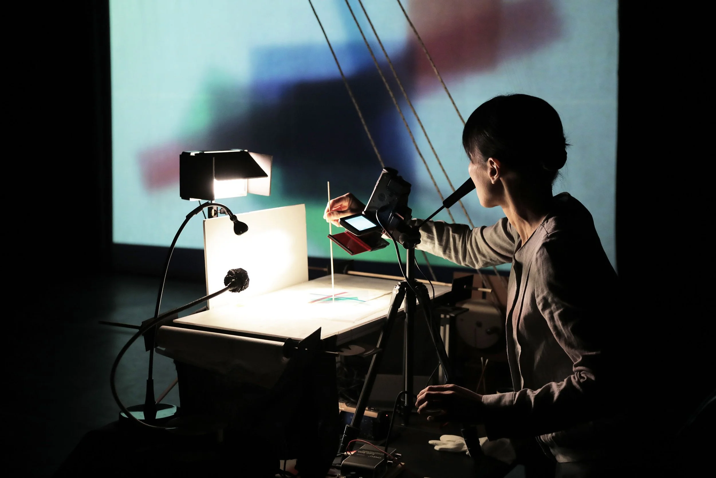 A person working on a desktop with various electronic equipment, illuminated by a small desk lamp, in front of a large screen displaying a blurry colorful image. The person is focused on their work and appears to be in a dark room.