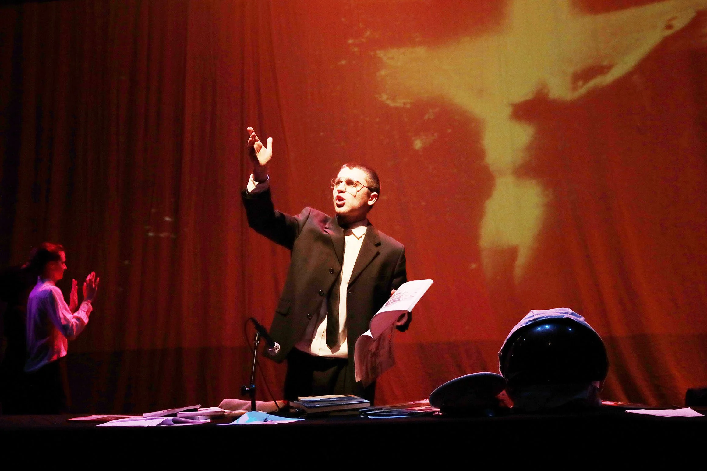 A man in a suit and glasses stands on stage, gesturing with his right hand, with a projection of his profile in yellow and orange behind him. There are women on the left clapping.