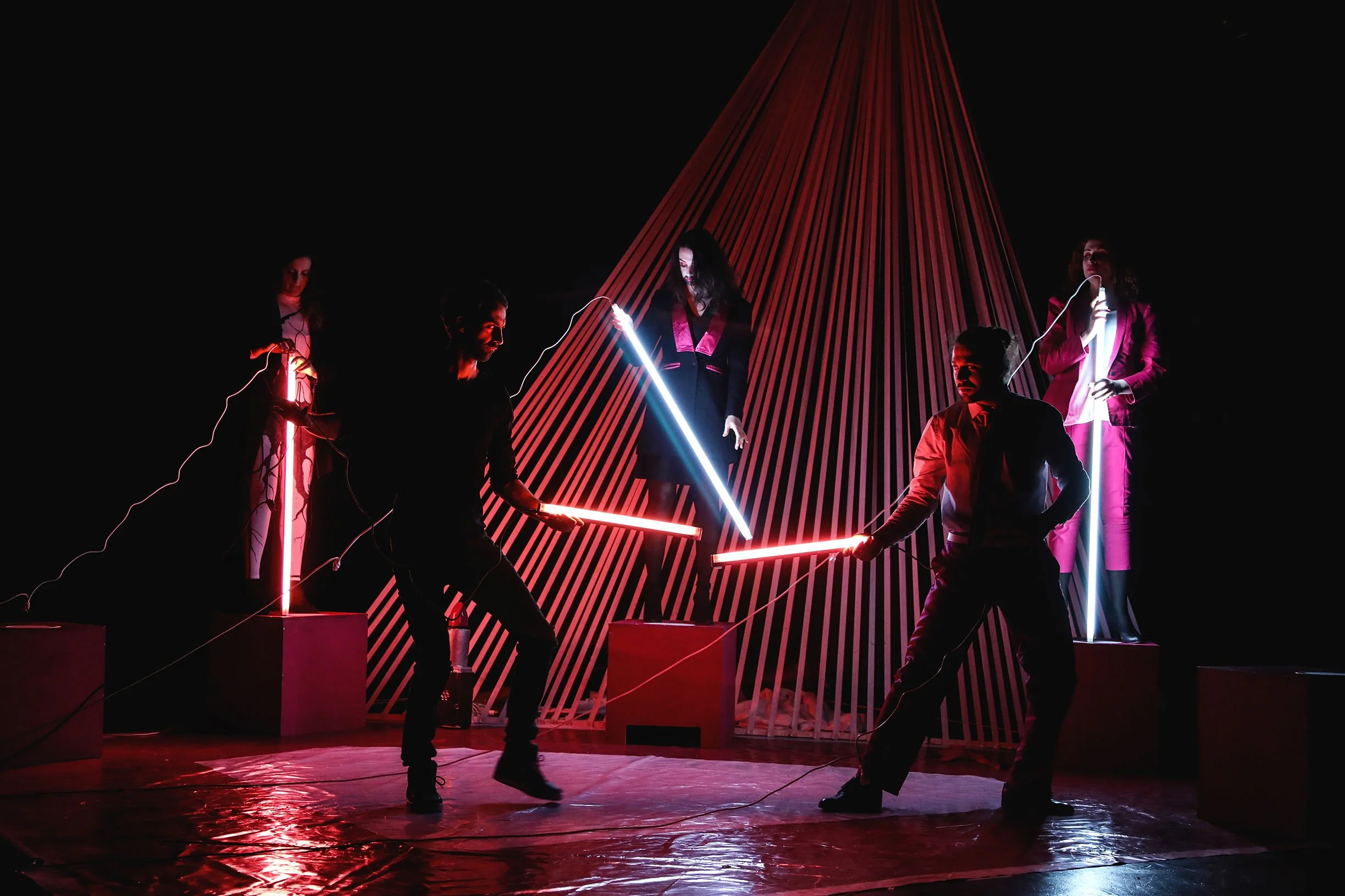 Six performers in dark clothing holding illuminated neon light tubes, two on each side, engaging in a dance or performance on a stage with geometric red and black striped backdrop, dim lighting, and reflective floor.