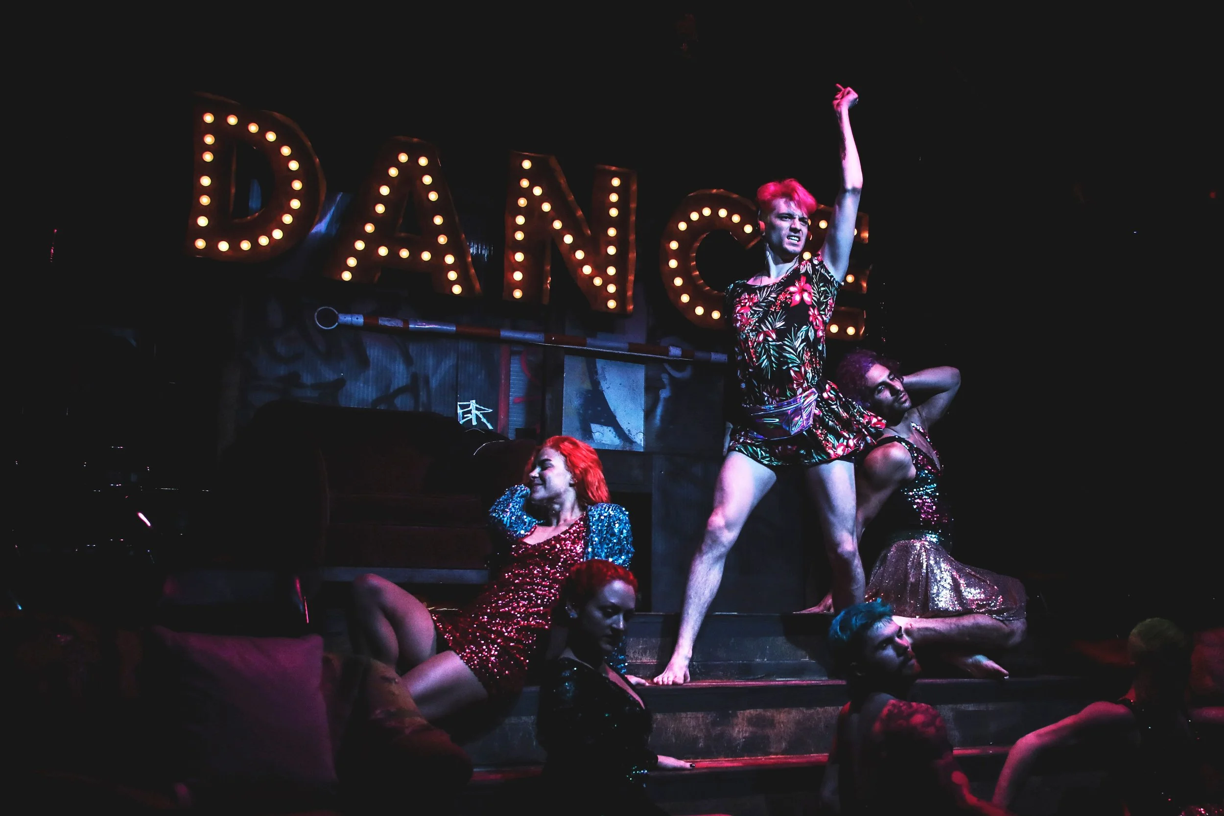 A theatrical performance featuring performers in colorful costumes, with a illuminated sign spelling 'DAN' in the background and performers striking dramatic poses.