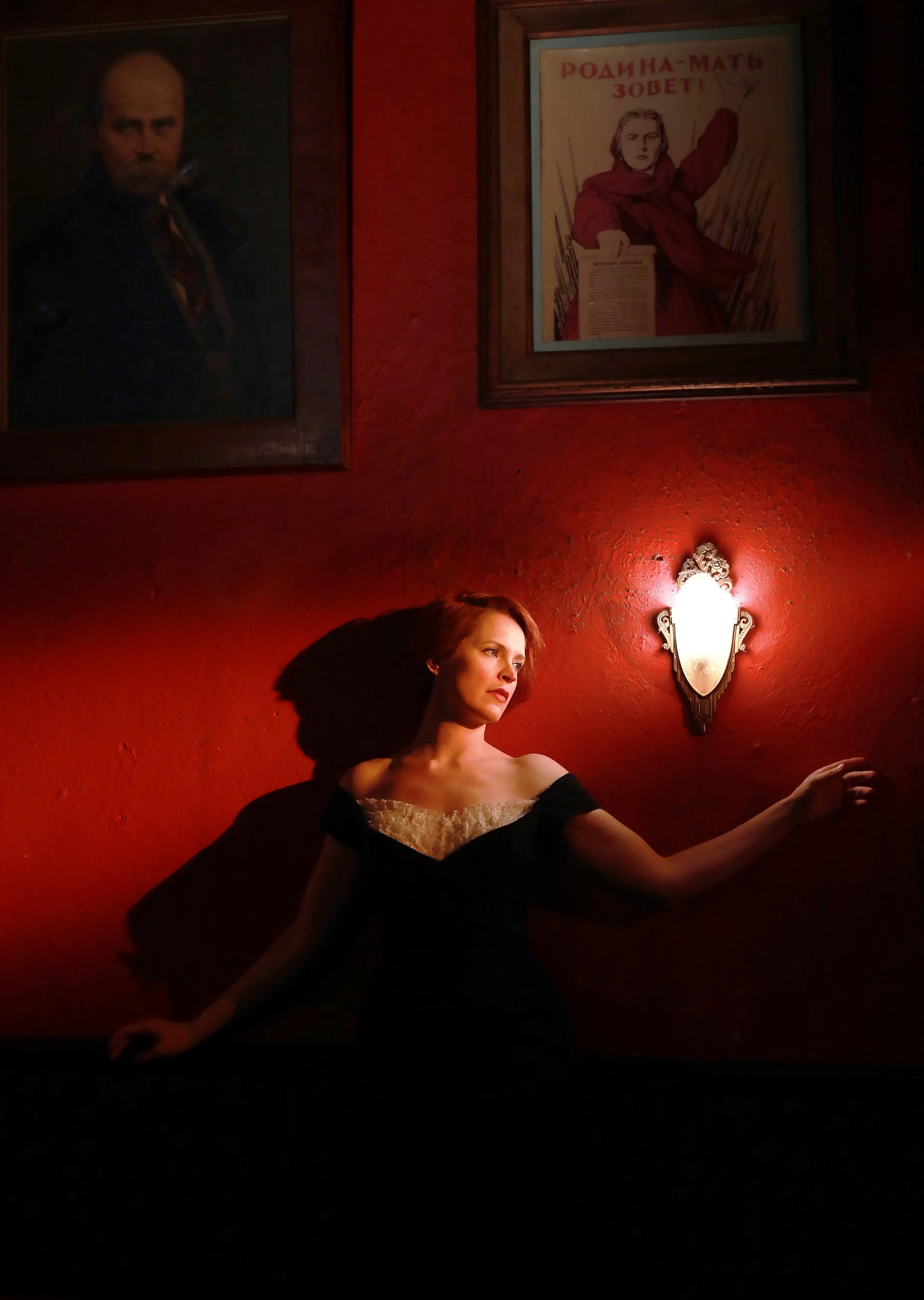 A woman with short brown hair sitting against a red wall illuminates from a wall-mounted light fixture. Behind her are framed posters on the wall, including a portrait of a man and a vintage poster with Russian text.