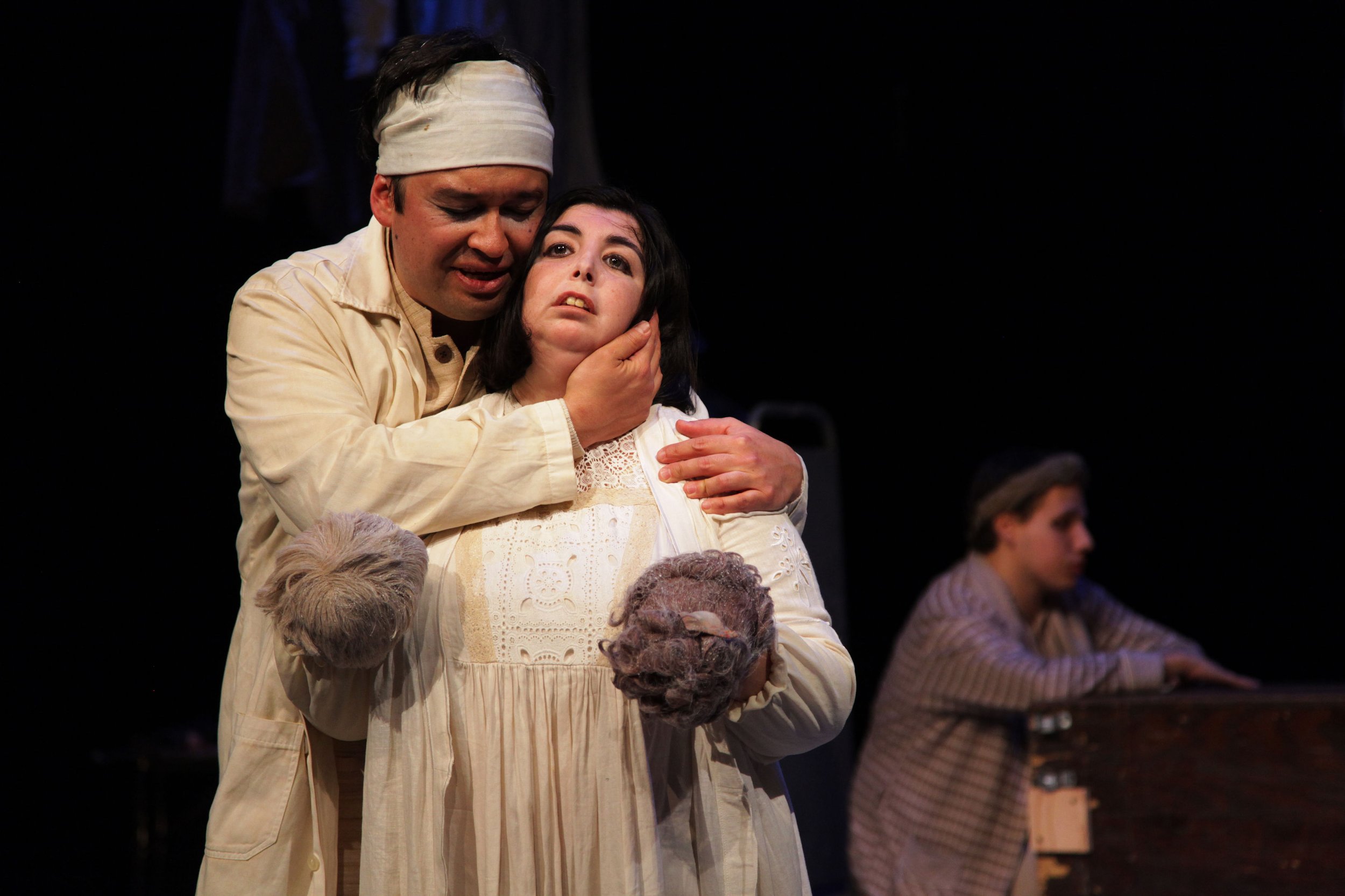 Two actors performing on stage, with one actor holding the face of the other actor, who appears distressed, amidst other cast members in the background.