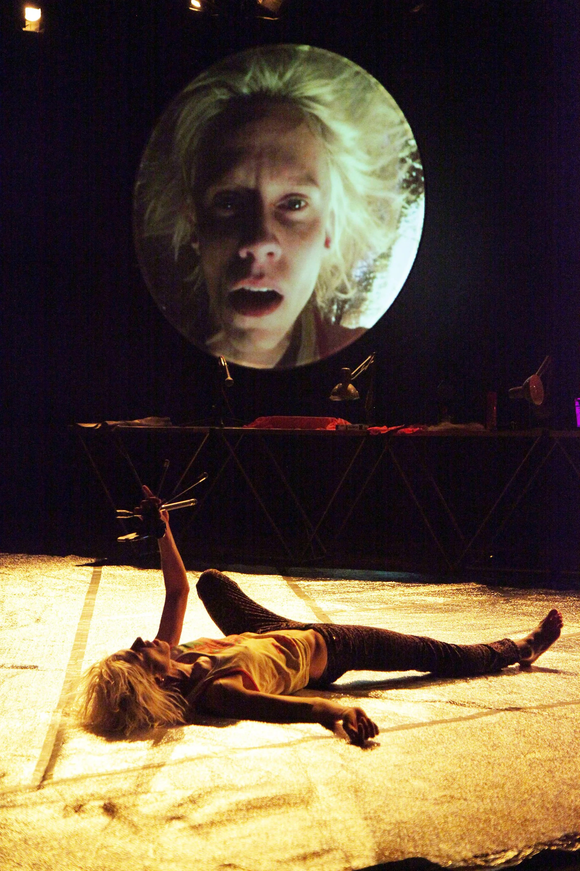 A person is lying on the stage floor with arms and legs extended, wearing black pants and a yellow shirt. Behind a large illuminated screen displaying a woman's face with an expression of surprise or fear.