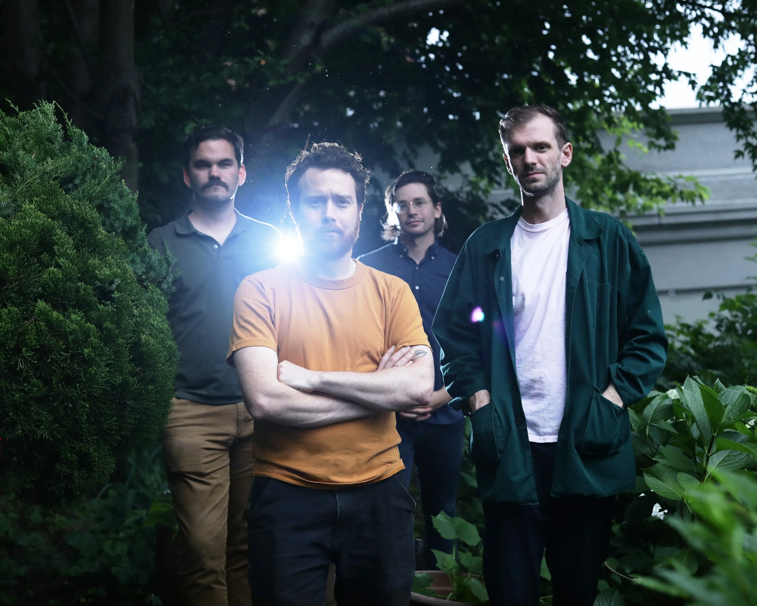 Four men standing in a lush garden or park, surrounded by green foliage, with the sunlight filtering through the trees.