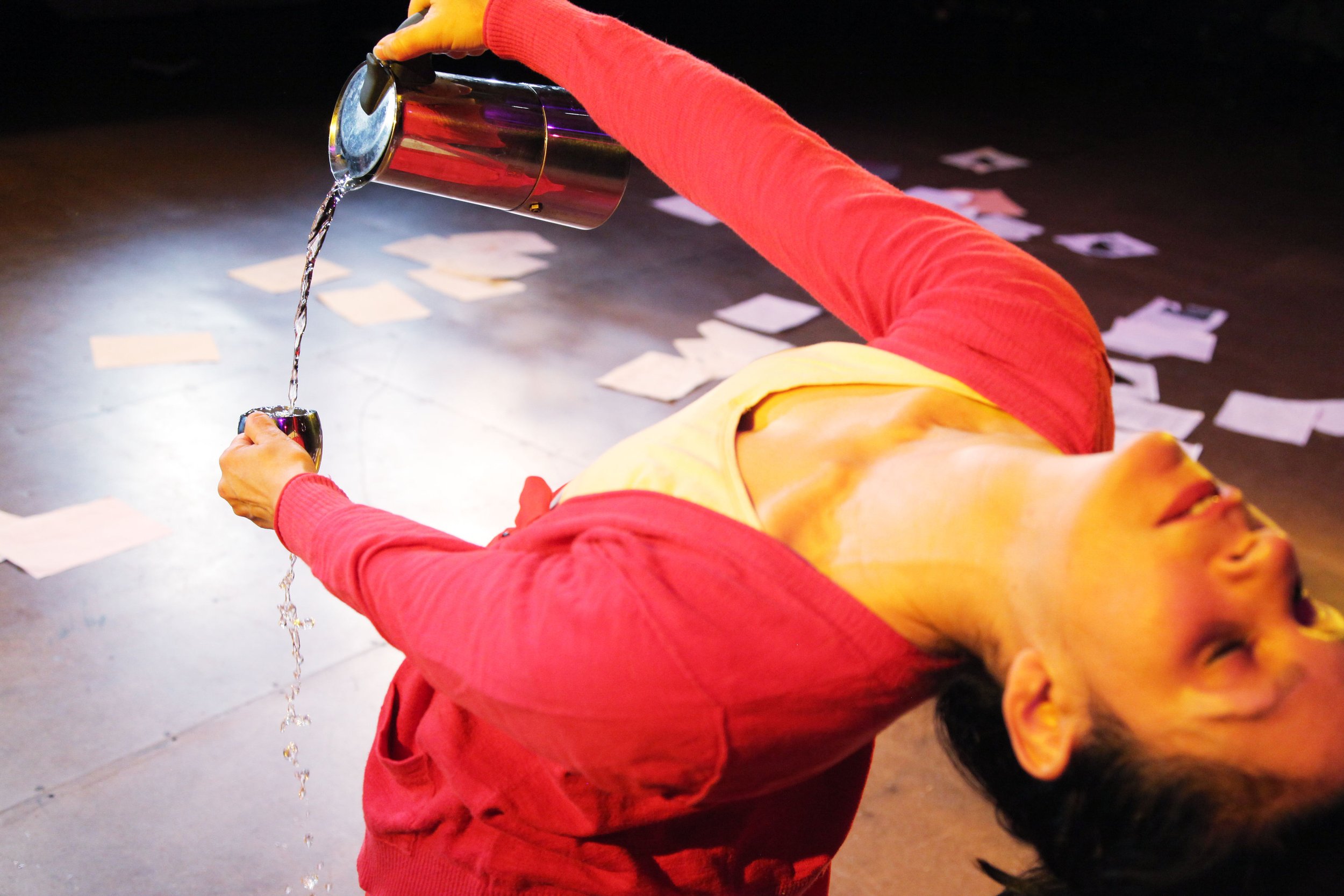 A person wearing a red long sleeve shirt and yellow pants, lying on their side on a dark surface, holding a shiny metal cup under a flowing stream of liquid from a metal can, with scattered pieces of paper on the surface.