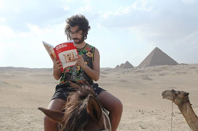 A man with curly hair and glasses sitting on a horse in a desert, reading a magazine, with pyramids visible in the background.