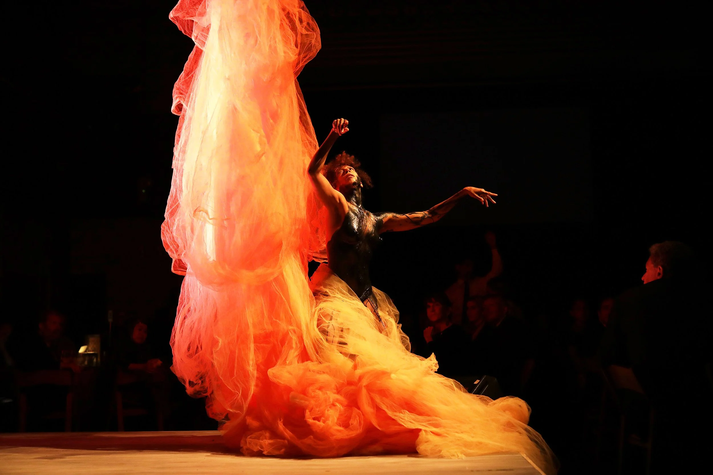 A person dancing on stage with flowing orange and yellow fabric, surrounded by an audience in the dark.