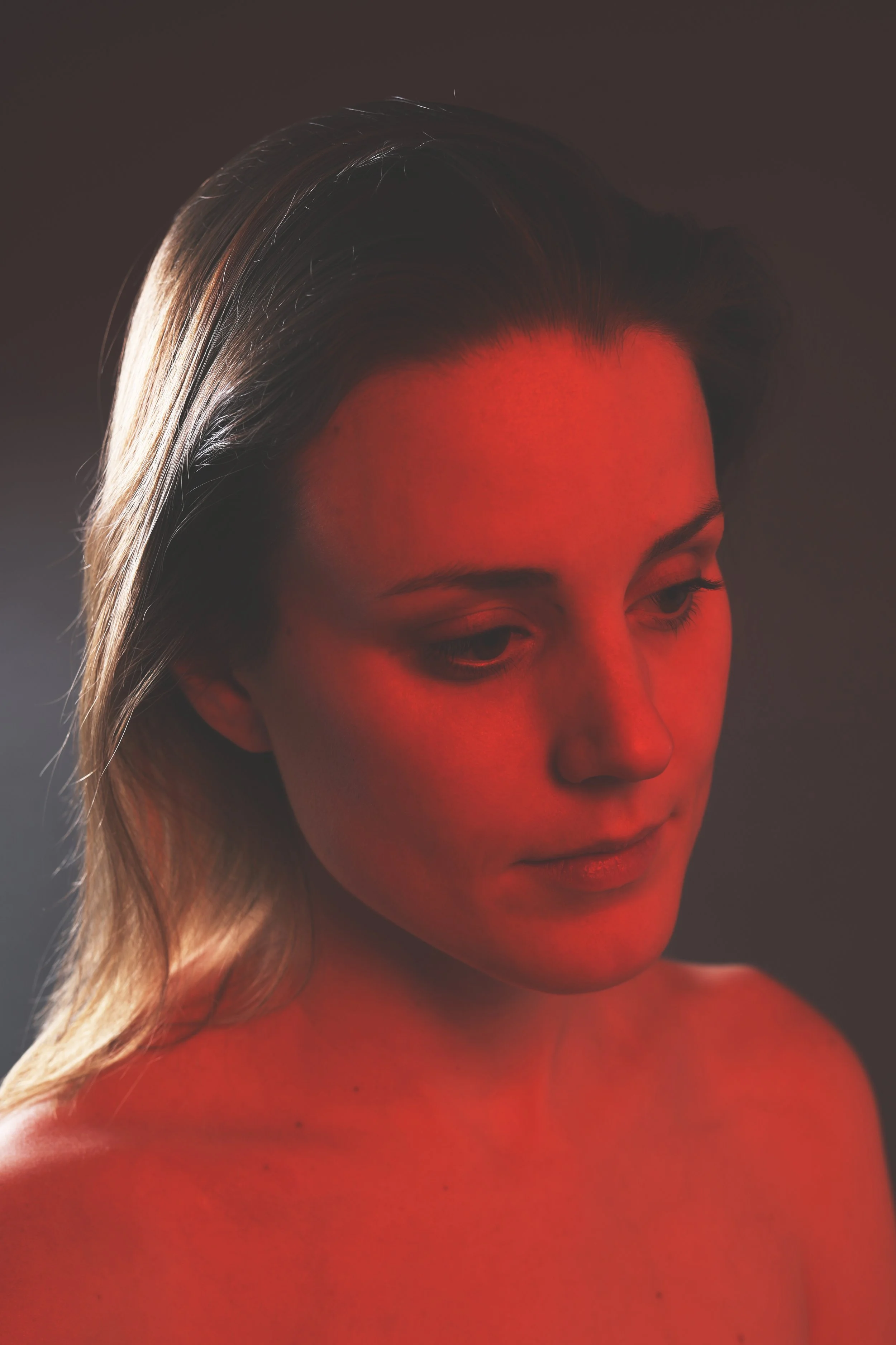 A woman with long blonde hair looking downward, with her face and upper chest illuminated by a red light, against a dark background.