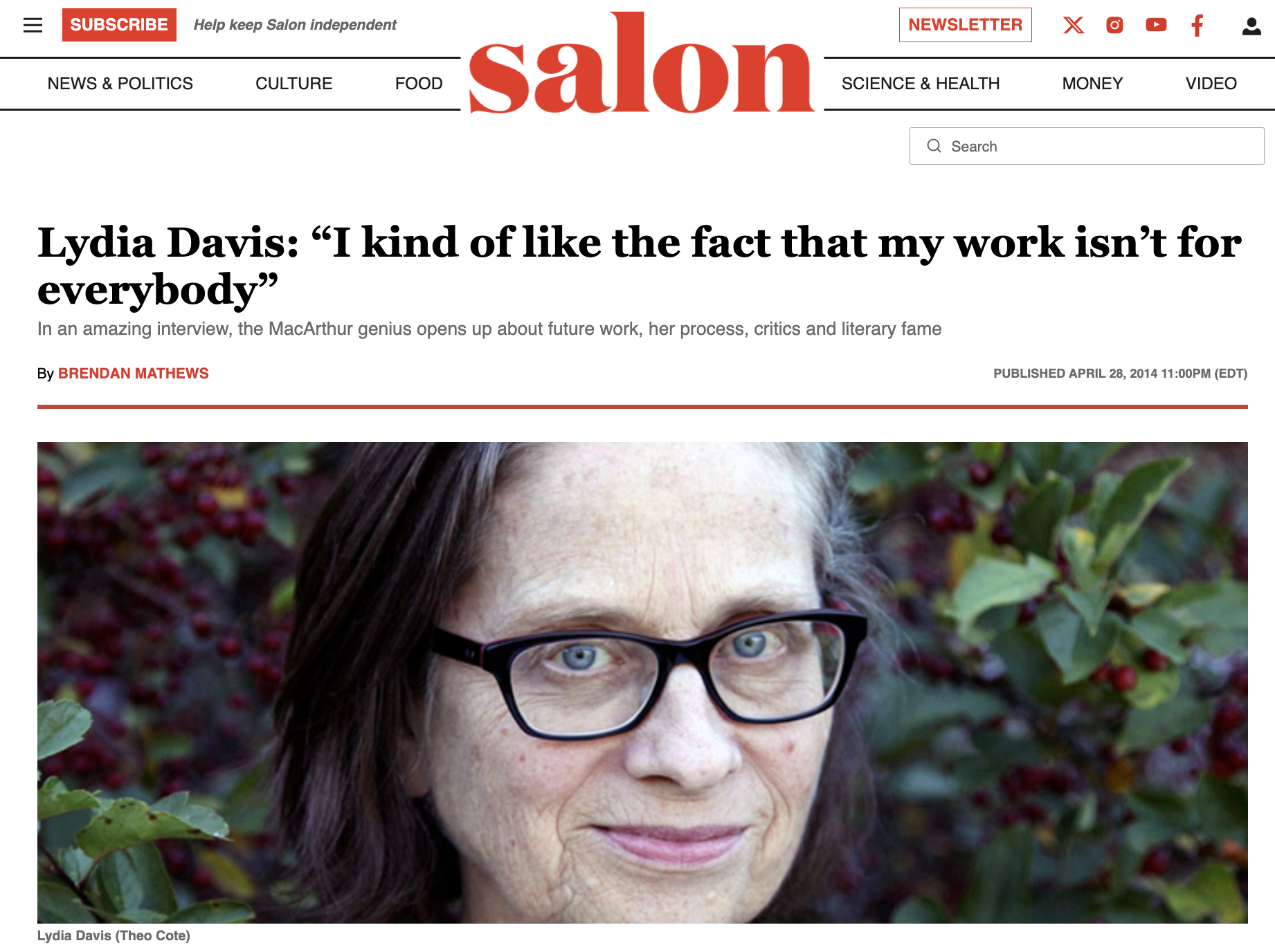 Close-up photo of Lydia Davis with blue eyes, wearing black glasses, surrounded by green and red foliage.