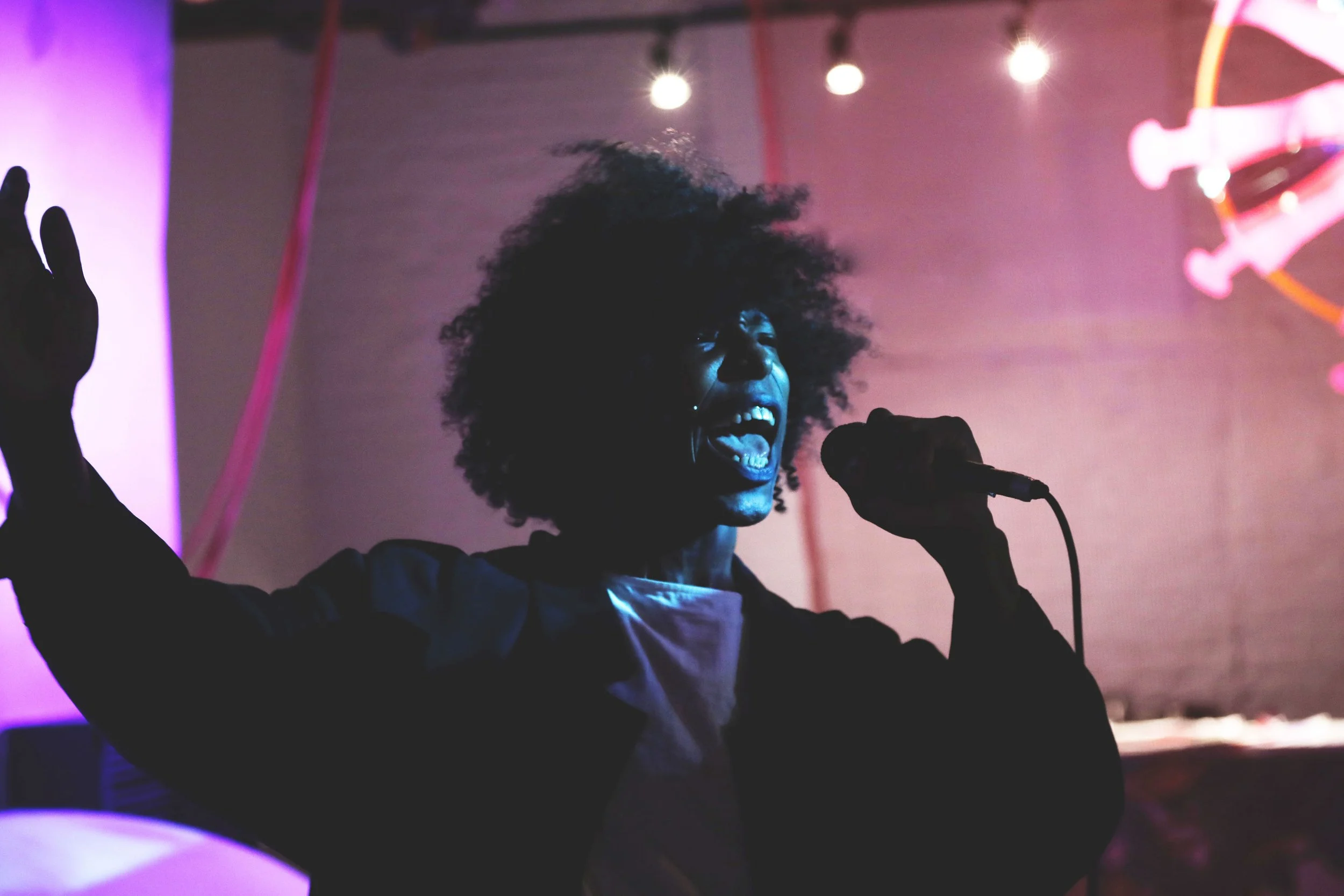 A person with curly hair singing or shouting into a microphone on stage, illuminated by colorful stage lights.