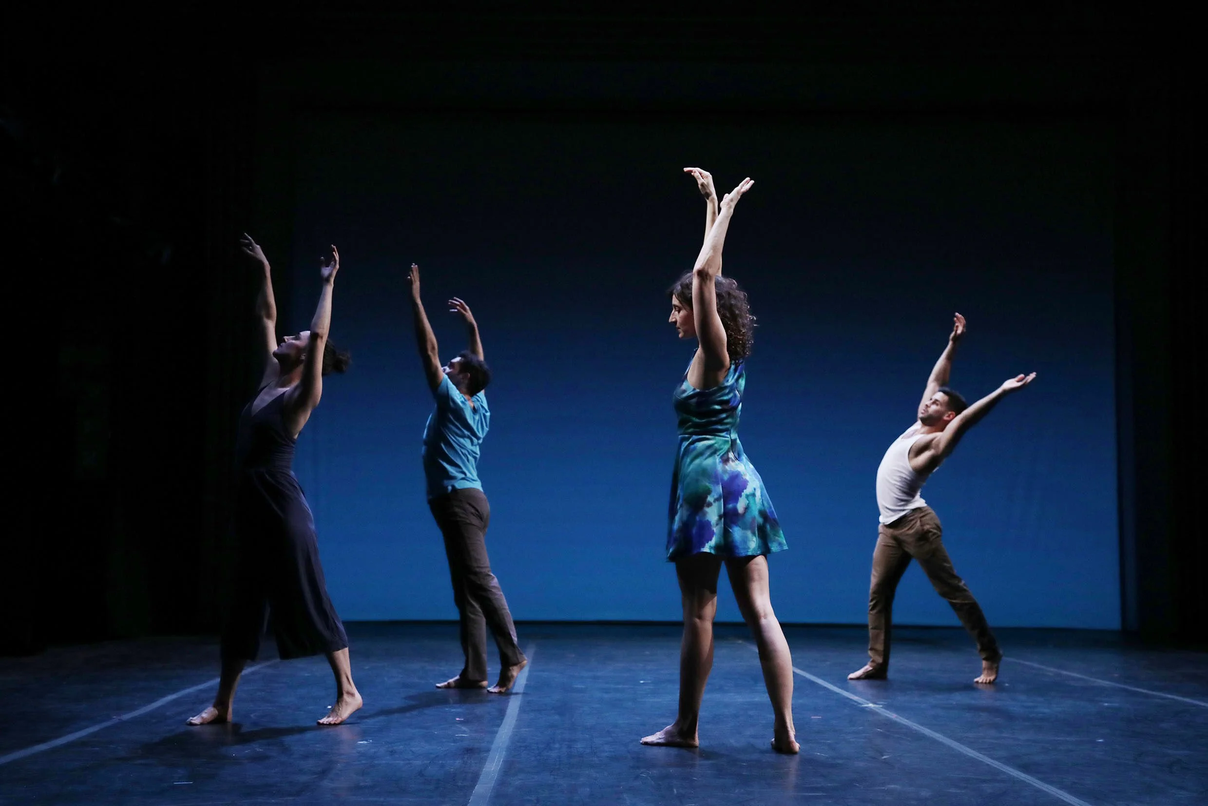 Group of dancers practicing on stage with arms raised in ballet poses.