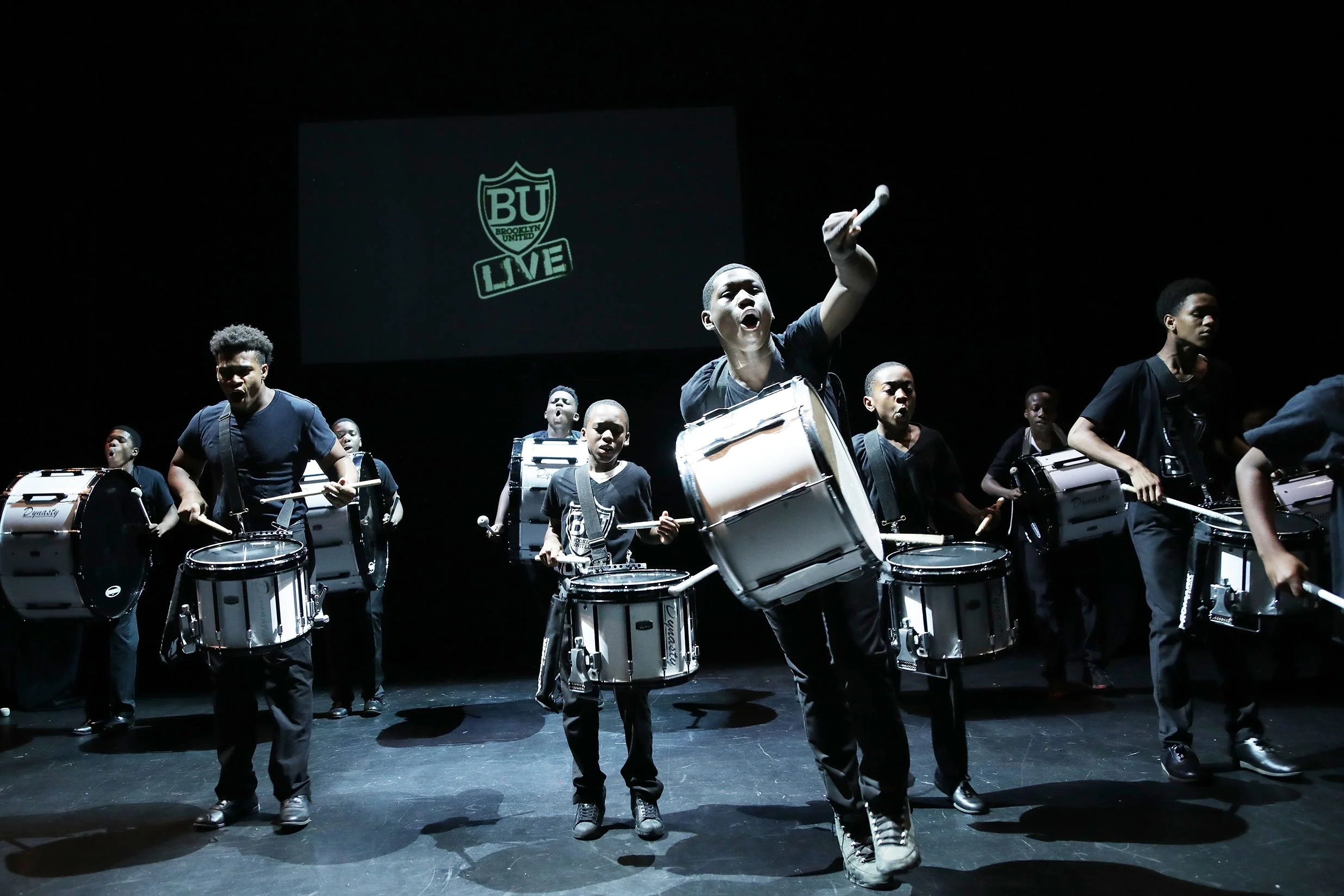A group of young boys playing drums on stage during a performance, with a screen in the background displaying the Brooklyn United logo and the word 'LOVE'.