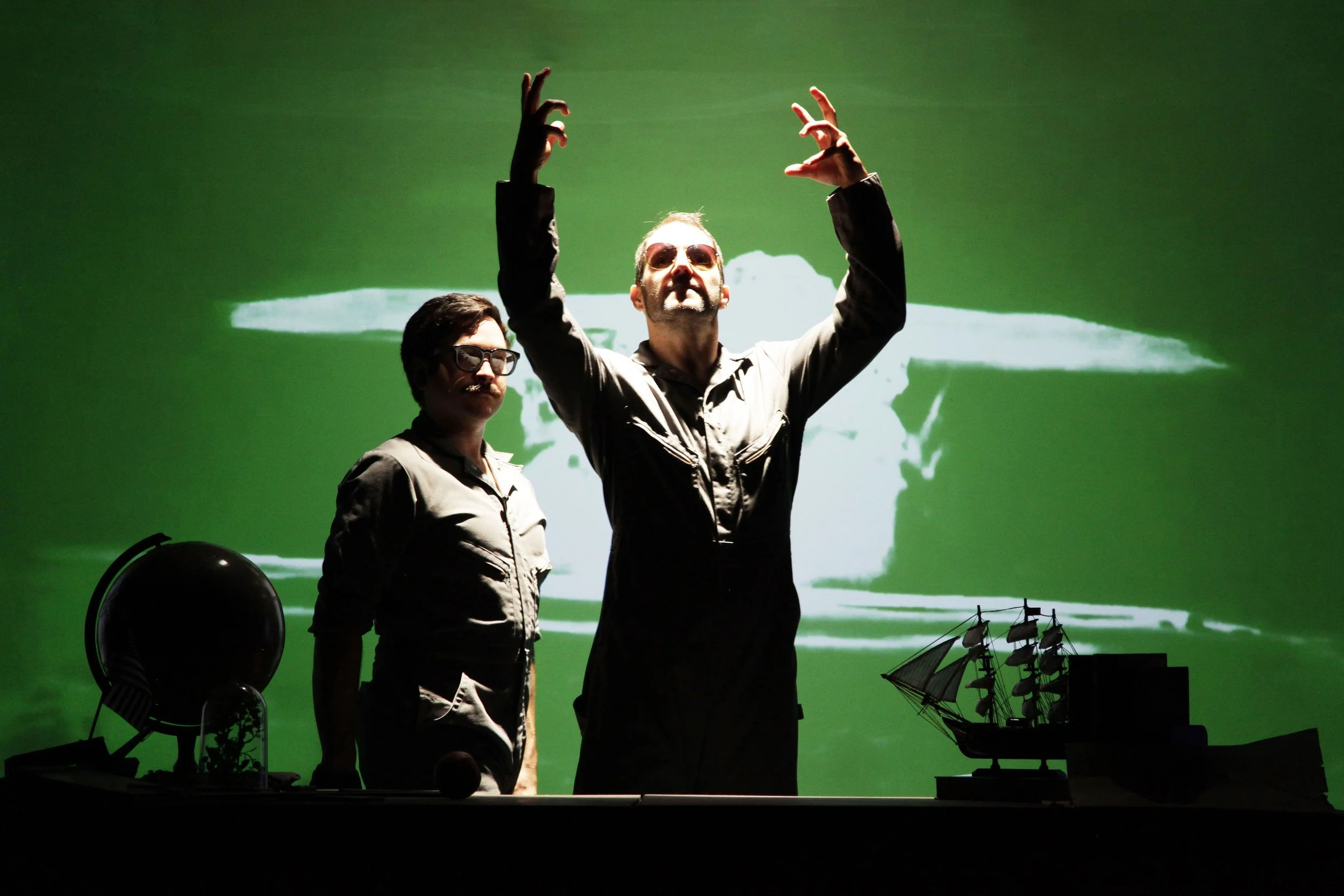 Two men in military-style clothing with sunglasses, one standing with hands raised in a dance or gesture, the other standing nearby with arms relaxed. Behind them is a green screen showing a naval scene with ships and sailing vessels, creating a hist