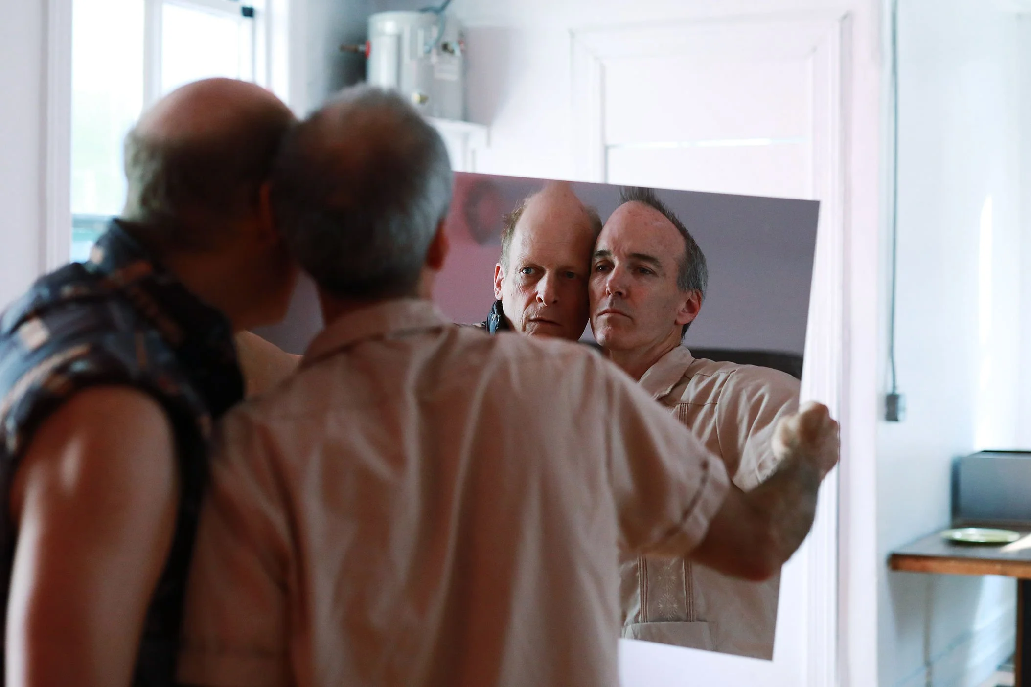 An elderly person looking at himself in a mirror with two younger people, possibly a woman and a man, in a home setting.
