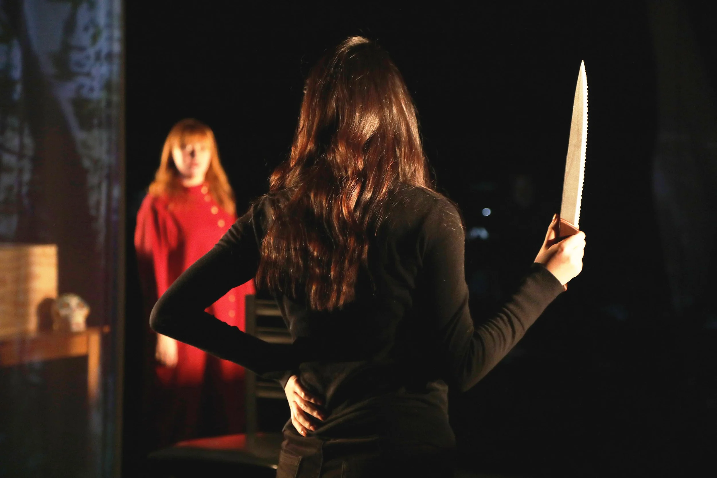 A woman with long dark hair holding a large kitchen knife in front of another woman with red hair dressed in red, on a dark stage with some background elements visible.