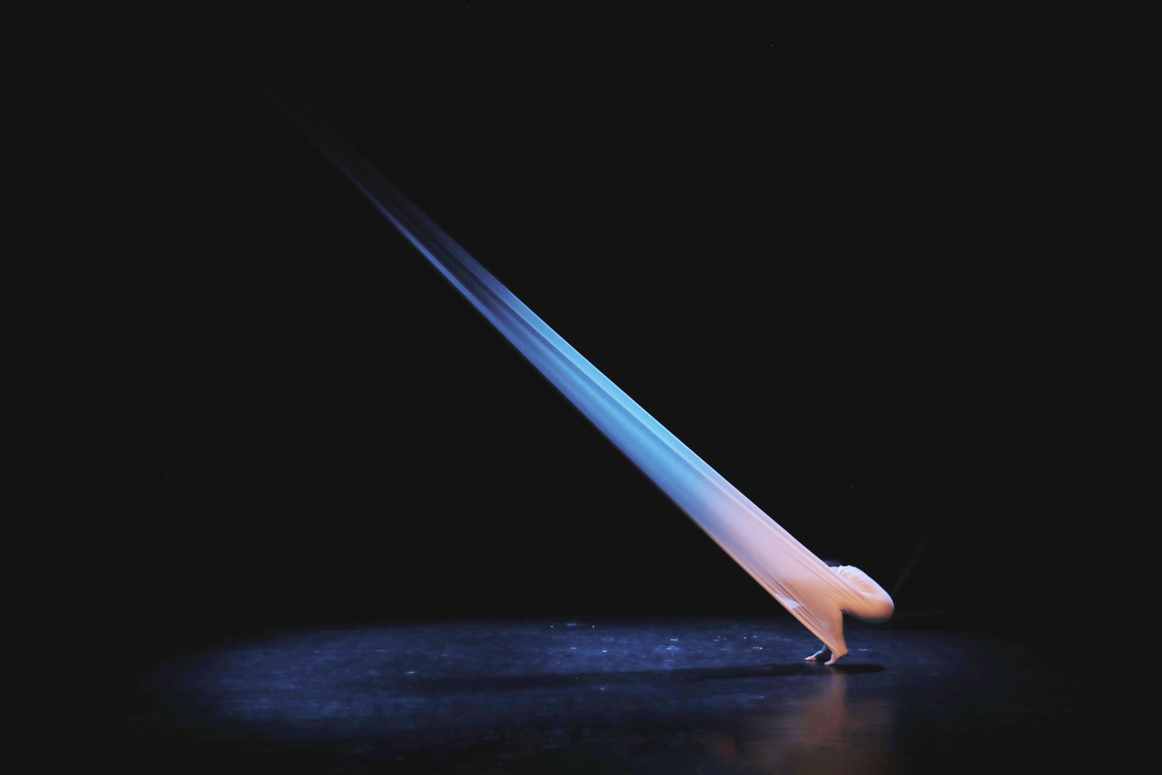 A performer in a white costume is balancing on their hands, with a large white fabric flowing diagonally across a dark stage. The fabric is illuminated by stage lighting, creating a dramatic visual effect.