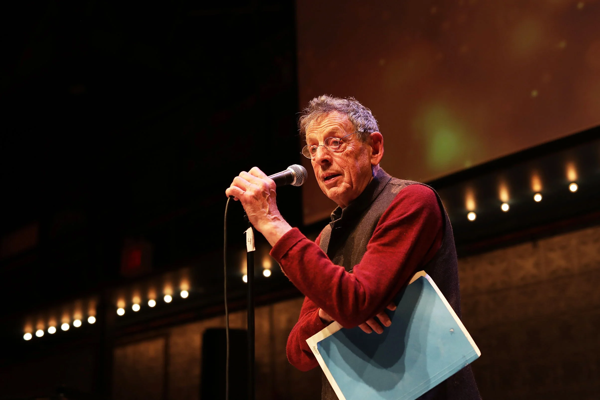 An elderly man with glasses speaking into a microphone at a public event, holding a large blue document or folder.