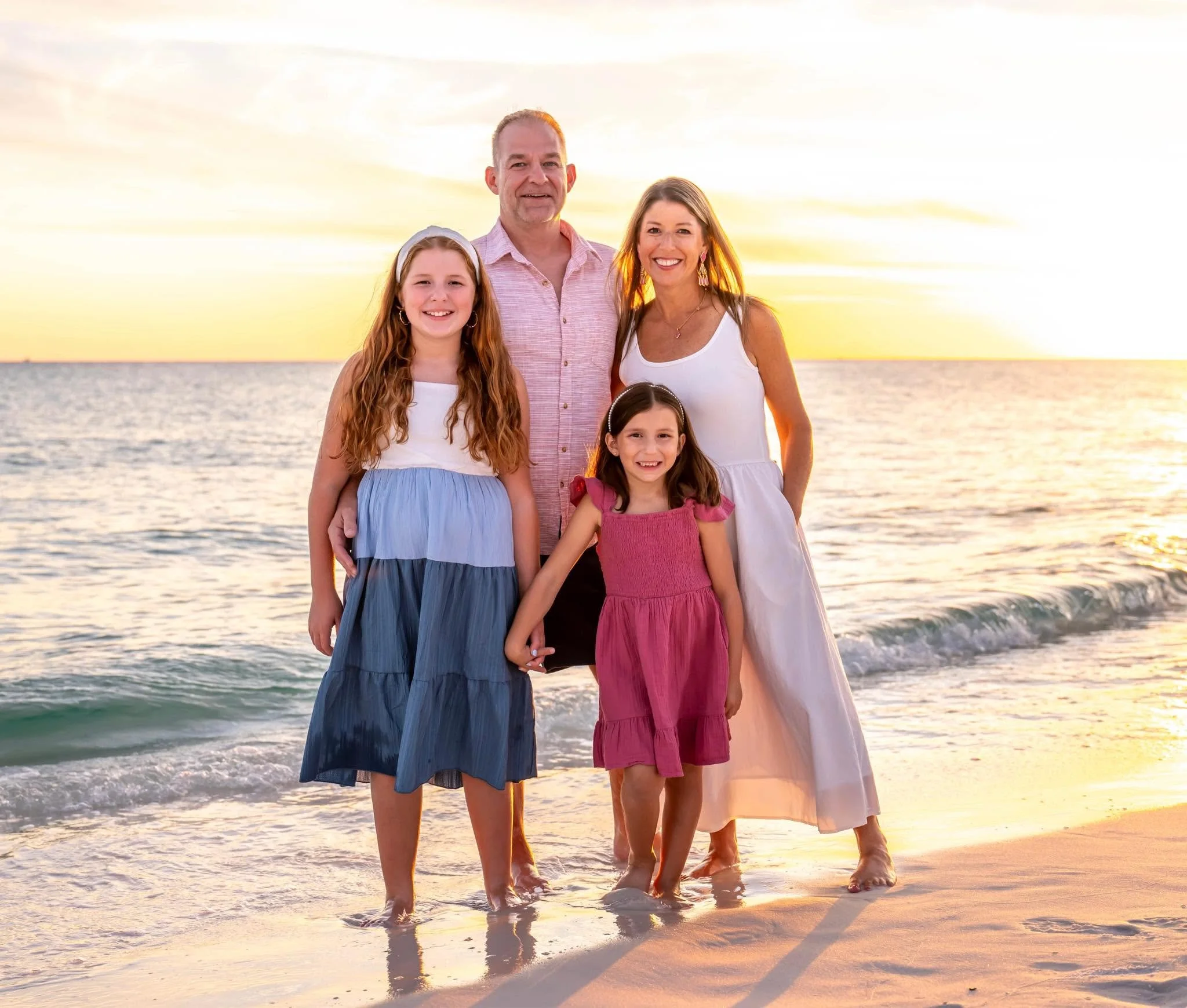 A family of four standing on a beach at sunset, with the ocean in the background, smiling and enjoying their time together.