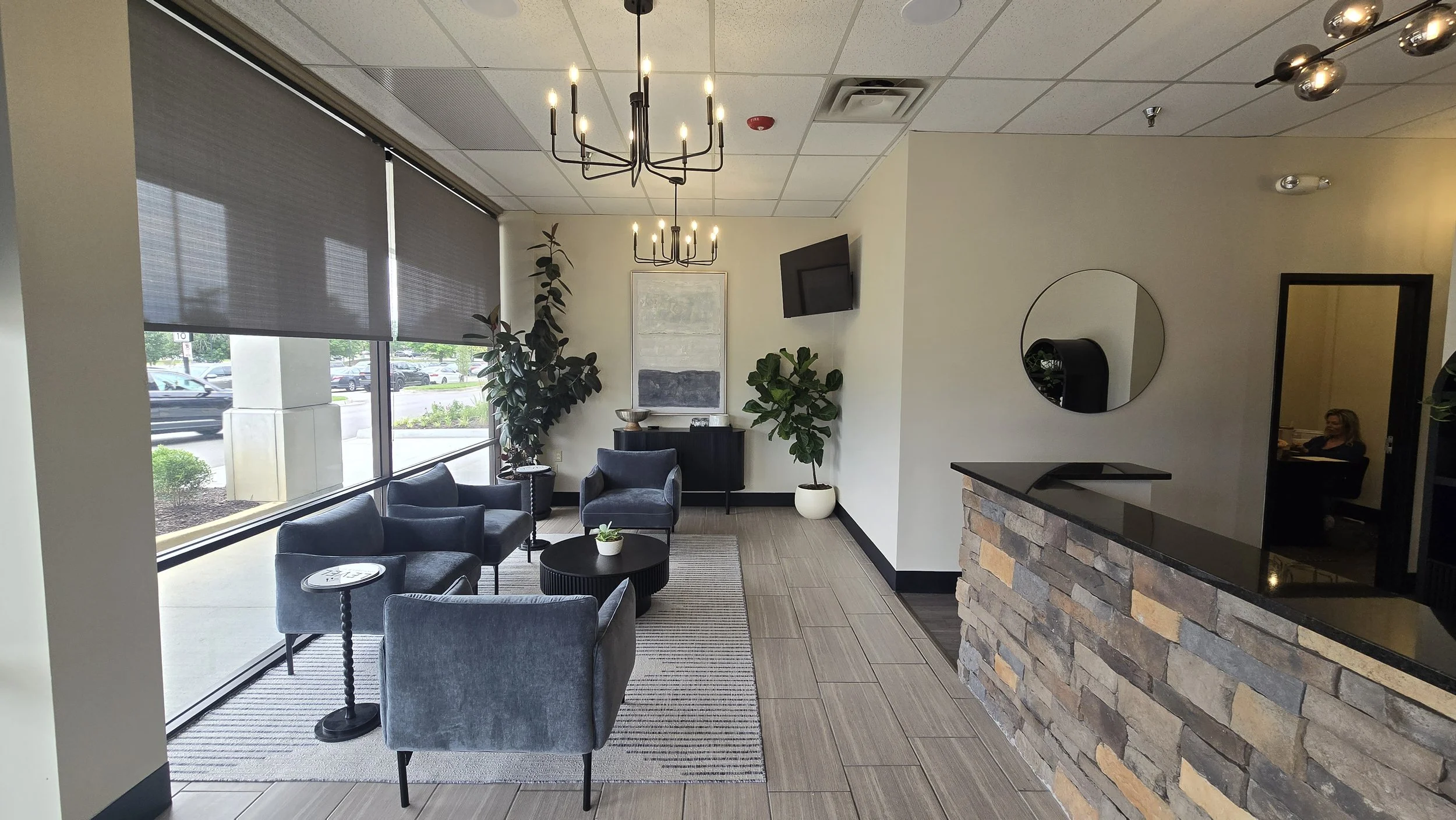 A modern indoor waiting area at Level Hormones and Health in South Overland Park. There are green plants, a painting, a wall-mounted TV, and a brick counter with a circular mirror.