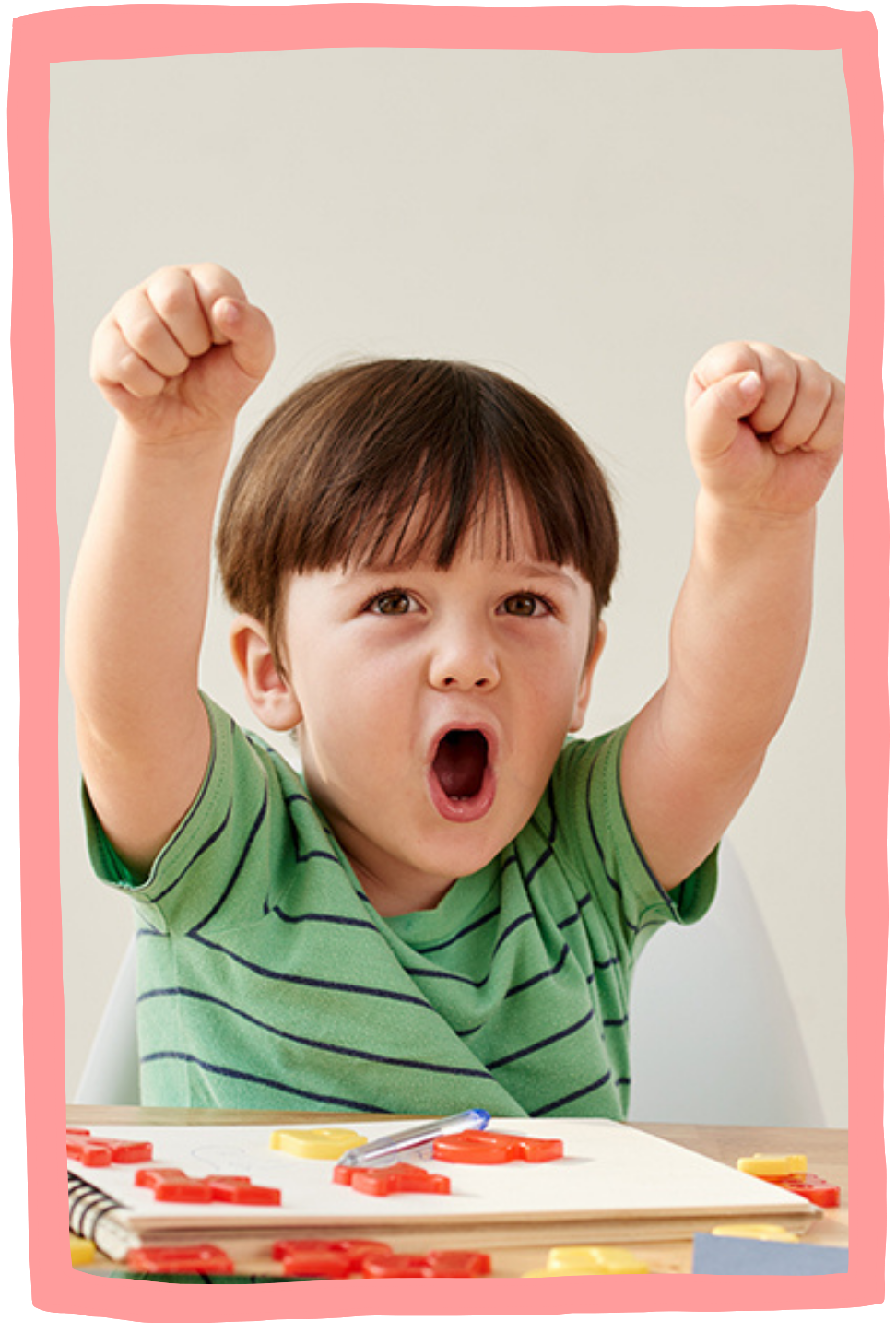 Young boy celebrating with raised fists and open mouth, playing with colorful letter tiles on a table during speech therapy for early language and literacy skills in New York City.