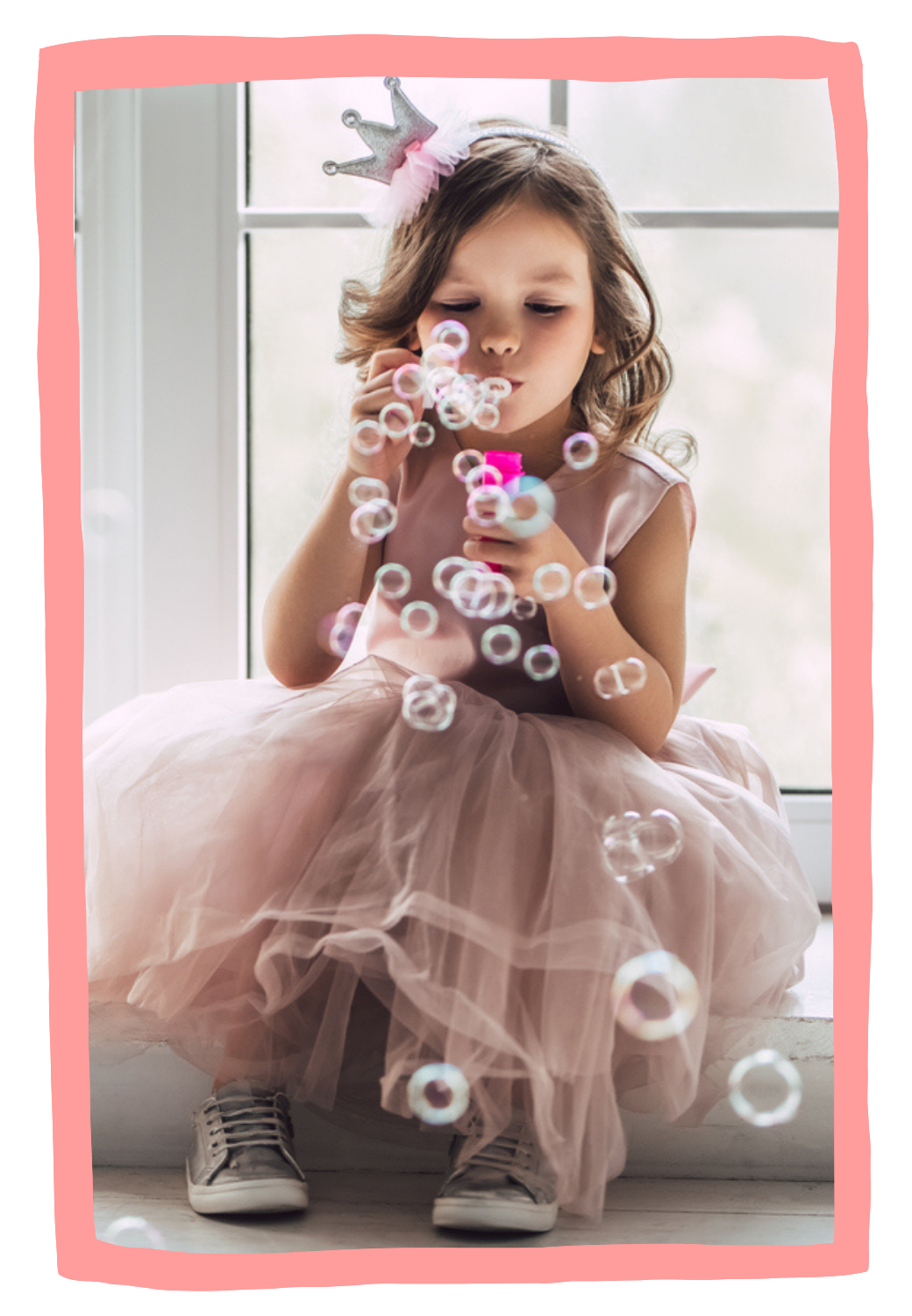 A young girl in a pink dress and sneakers sitting by a window, blowing bubbles with a bubble wand, wearing a crown headband, working on speech and language skills during in-home speech therapy for kids in New York City