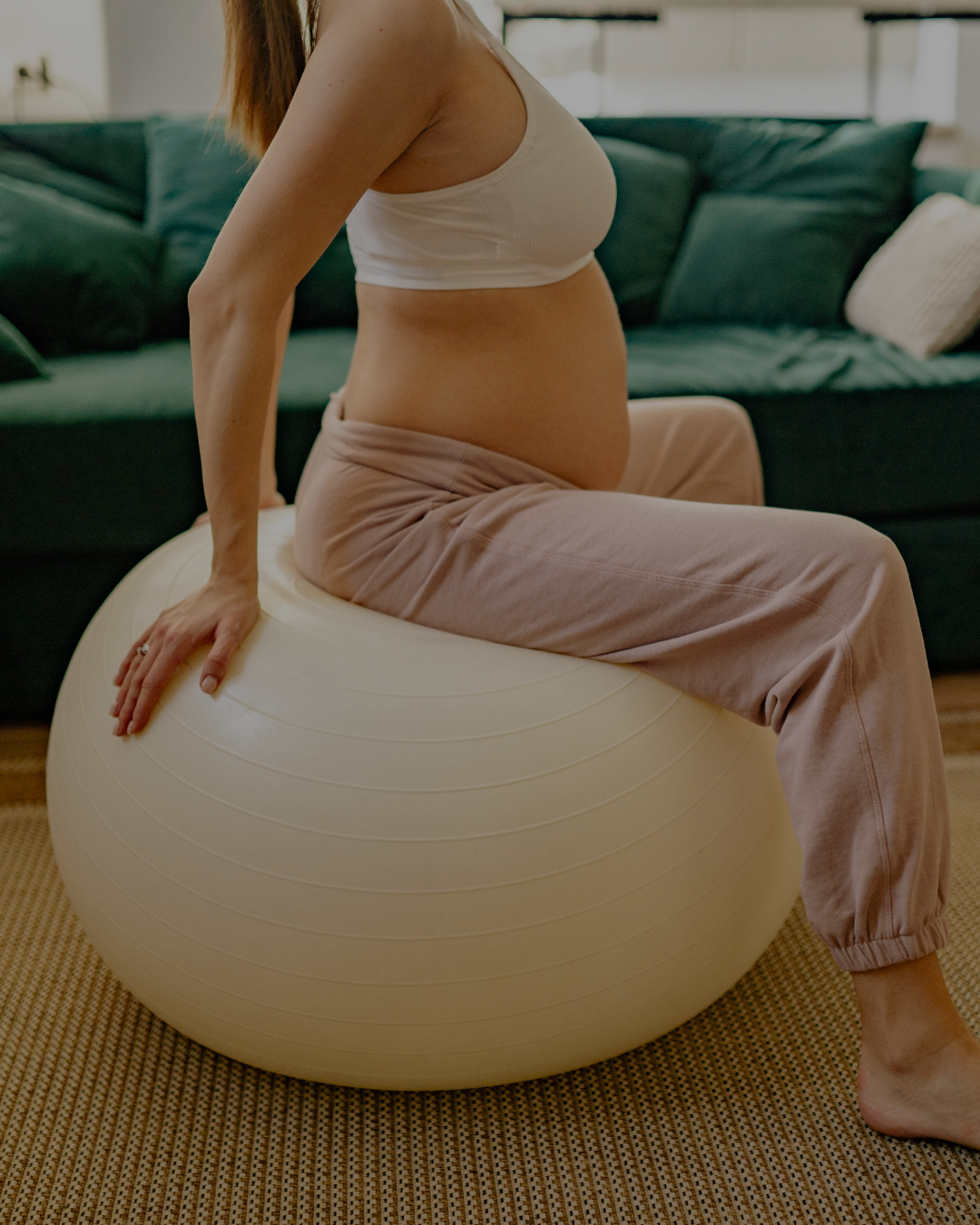 A pregnant woman sitting on an exercise ball in a living room, wearing a white sports bra and beige sweatpants.