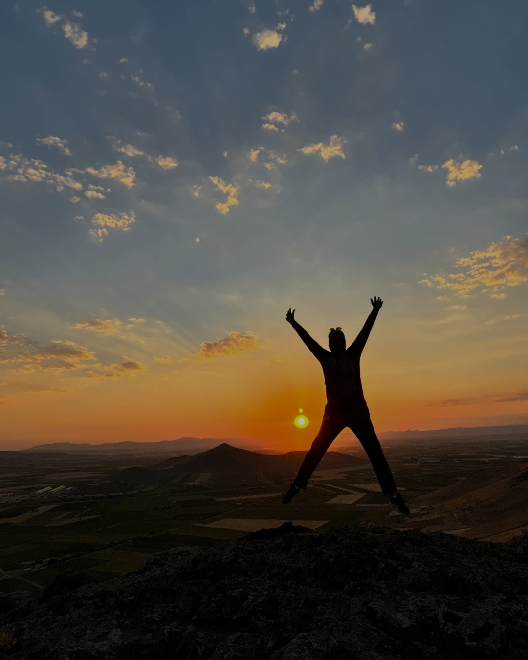 Person jumping on a rocky hilltop during a sunset with a colorful sky and scattered clouds in the background.