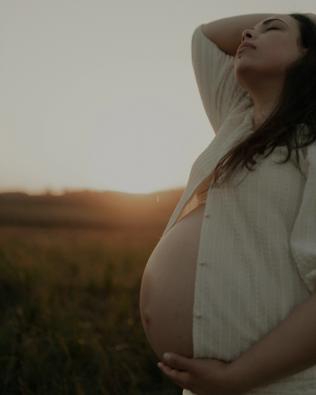 Pregnant woman standing outdoors during sunset, with her hand on her belly and eyes closed, in a relaxed pose.