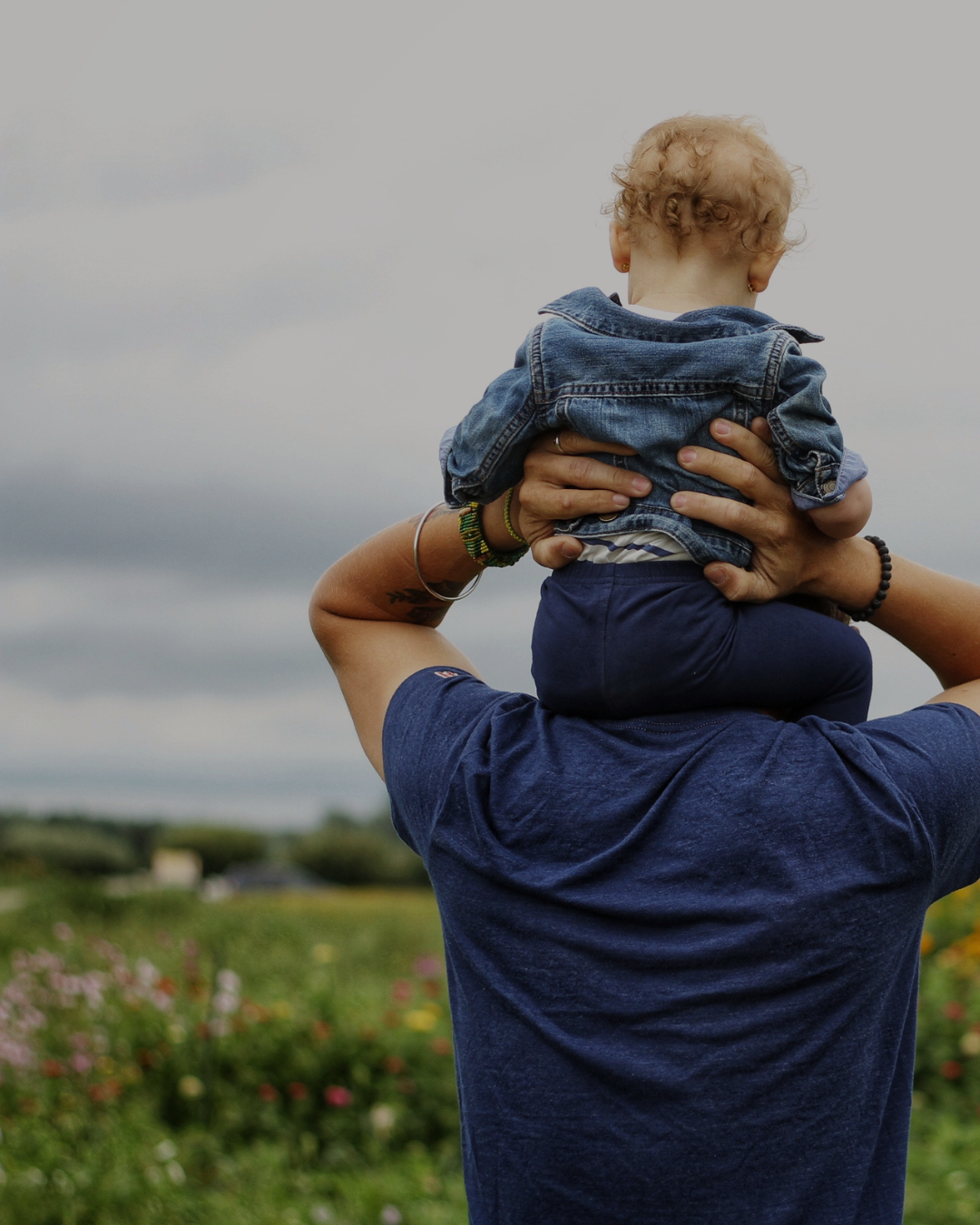 A person is holding a young child on their shoulders in an outdoor setting with a cloudy sky and greenery in the background.