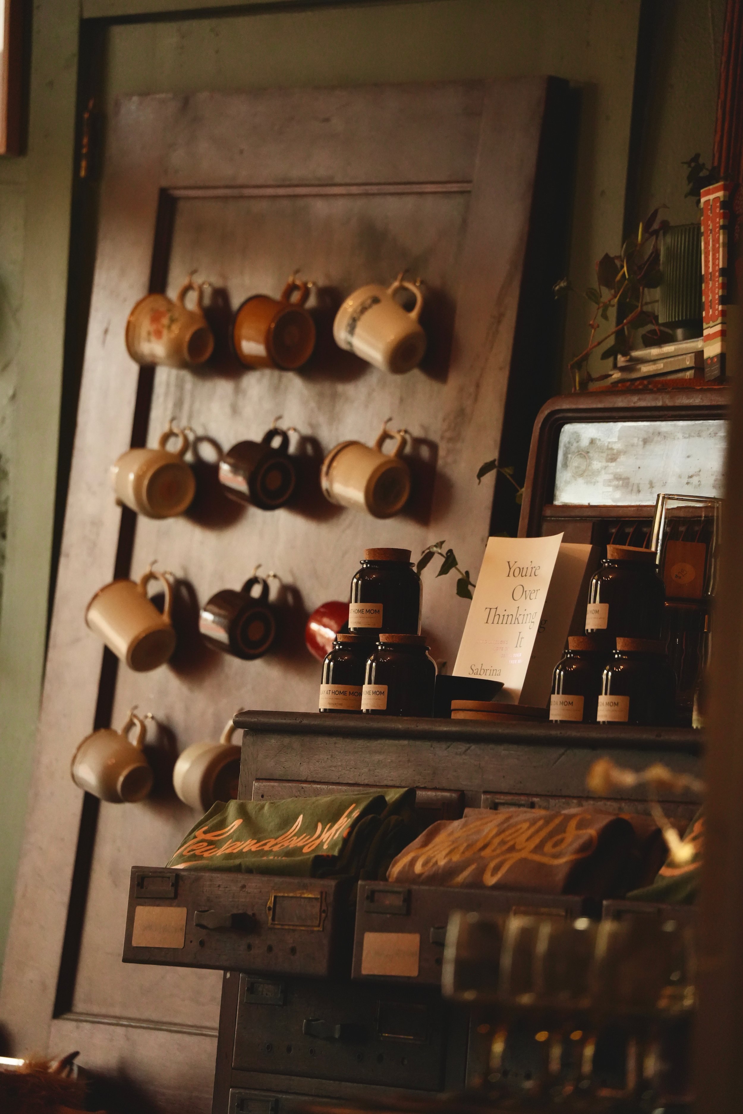 A wall with multiple mugs hanging on hooks and shelves with various items including bottles, a mirror, and a plant, in a cozy room.