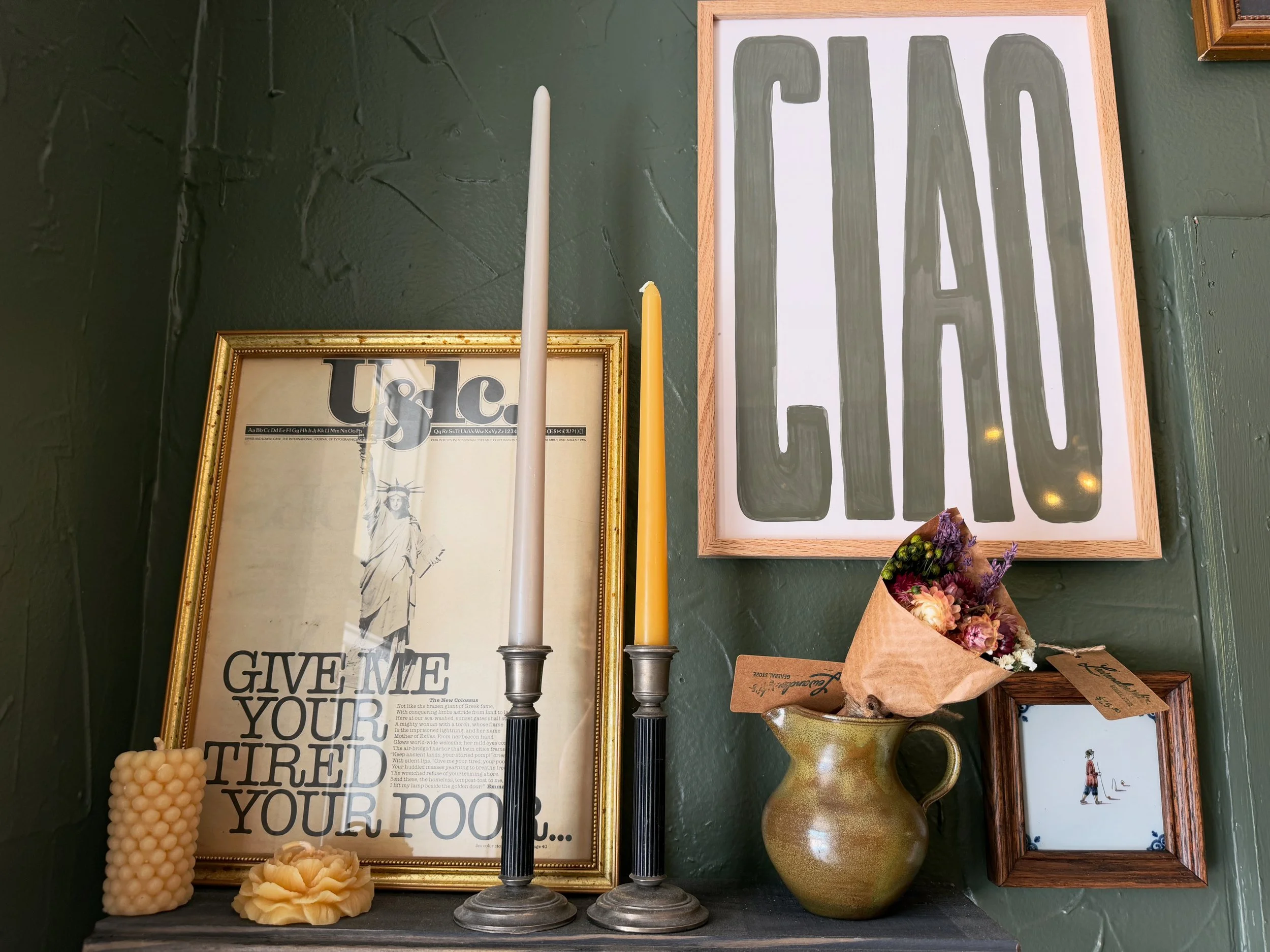 Decorative wall display featuring framed art, candles, a bouquet of dried flowers, and a poster with large text and a drawing of the Statue of Liberty.