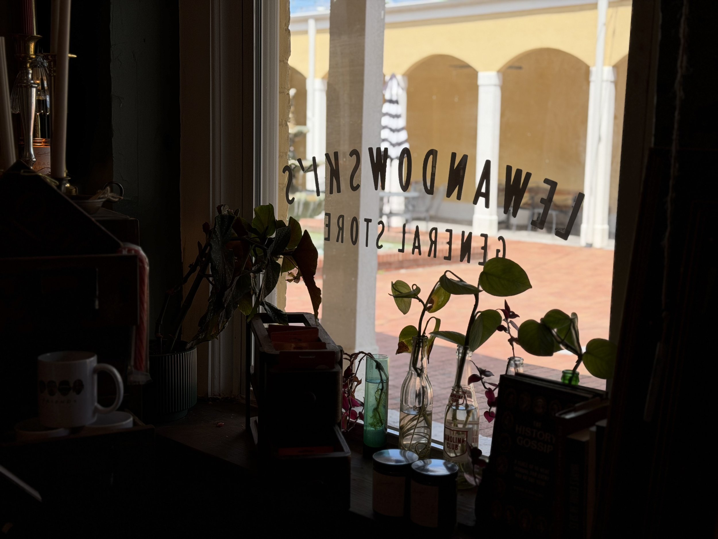 View from inside a cafe or shop looking out the window with plants and bottles on the windowsill, and a yellow building with arches outside.