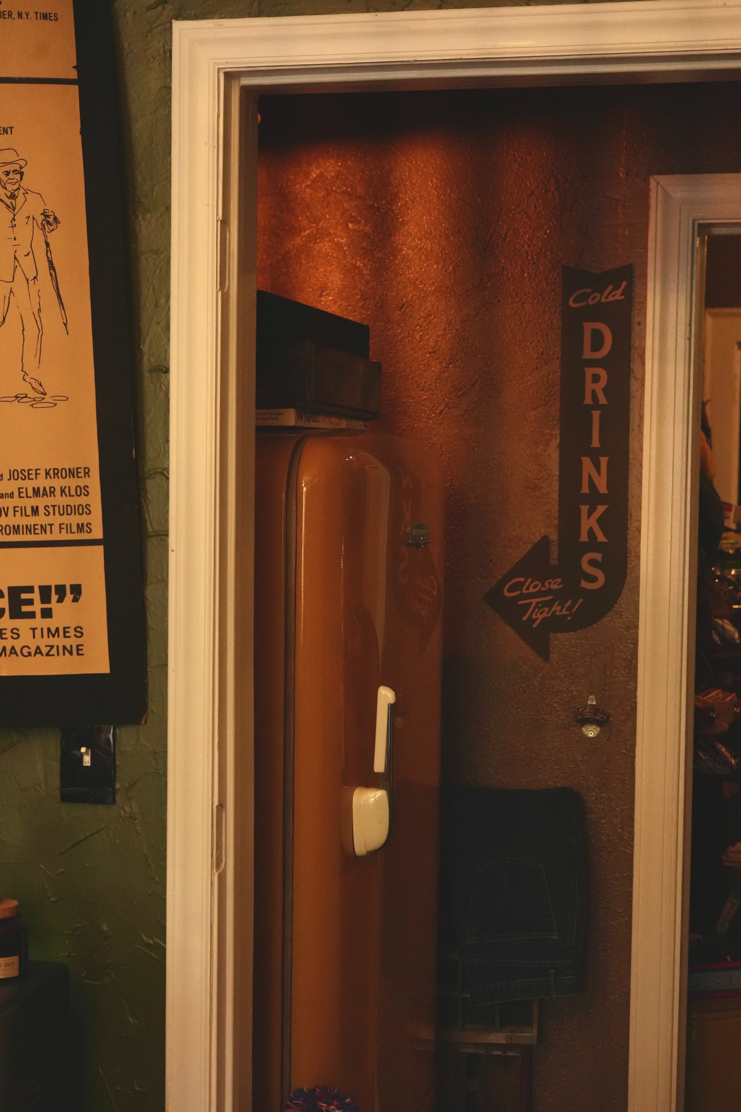 Interior view of a door opening into a dimly lit room with a textured wall, a sign that reads 'Cold Drinks, Close Tights!', and some furniture including a table and chairs.