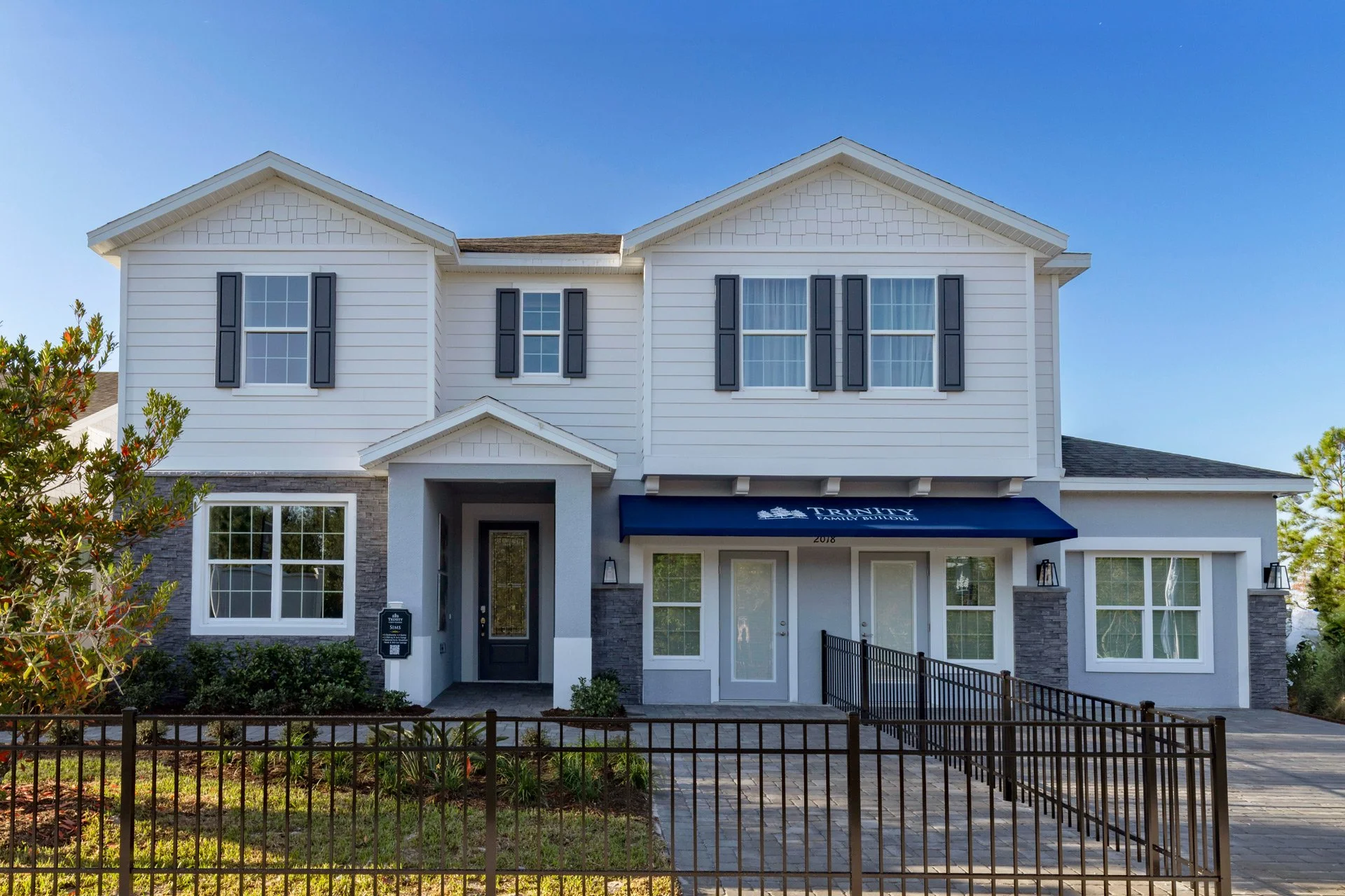 Front view of a modern, two-story house with white siding, black window shutters, and a black front door. The house has a small front yard with a black metal fence and some bushes. There is a blue awning over the entrance with illegible white text.