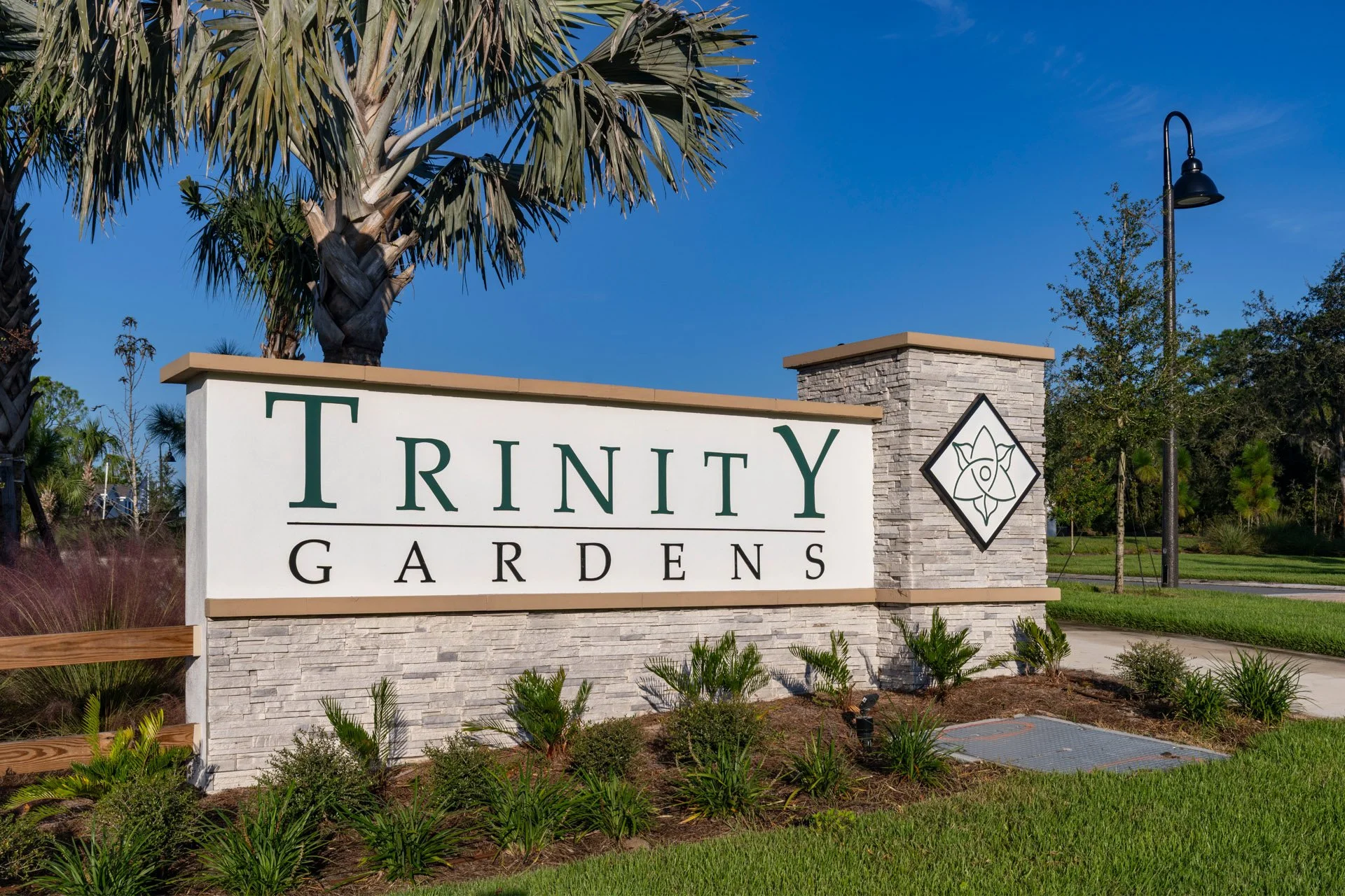 Entrance sign for Trinity Gardens with stone pillars, trees, landscaping, and clear blue sky in the background.