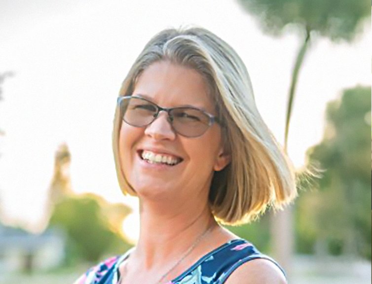A middle-aged woman smiles outdoors, wearing glasses and a patterned sleeveless top, with a blurred natural background.