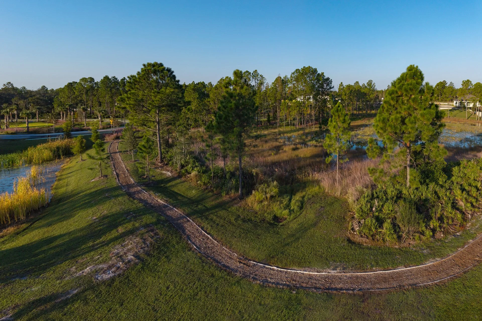 Aerial view of a landscape with trees, a small pond, and a walking trail. There's a clear blue sky above.