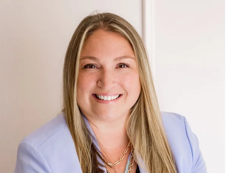 A woman with long blonde hair smiling, wearing a light blue blazer and layered necklaces, standing in front of a light-colored wall.